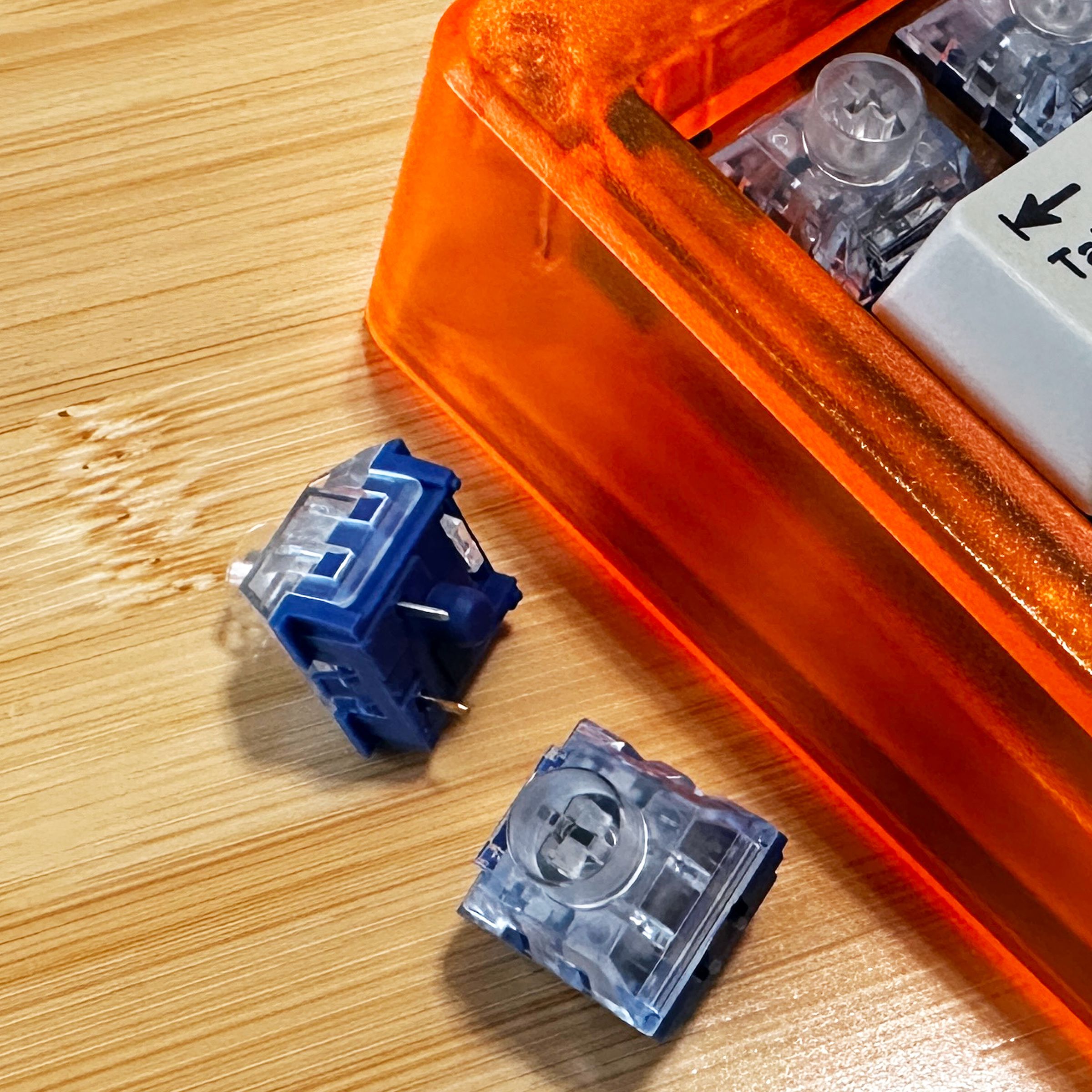 Close up of mechanical keyboard switches with clear tops and blue bases next to a bright orange keyboard. 