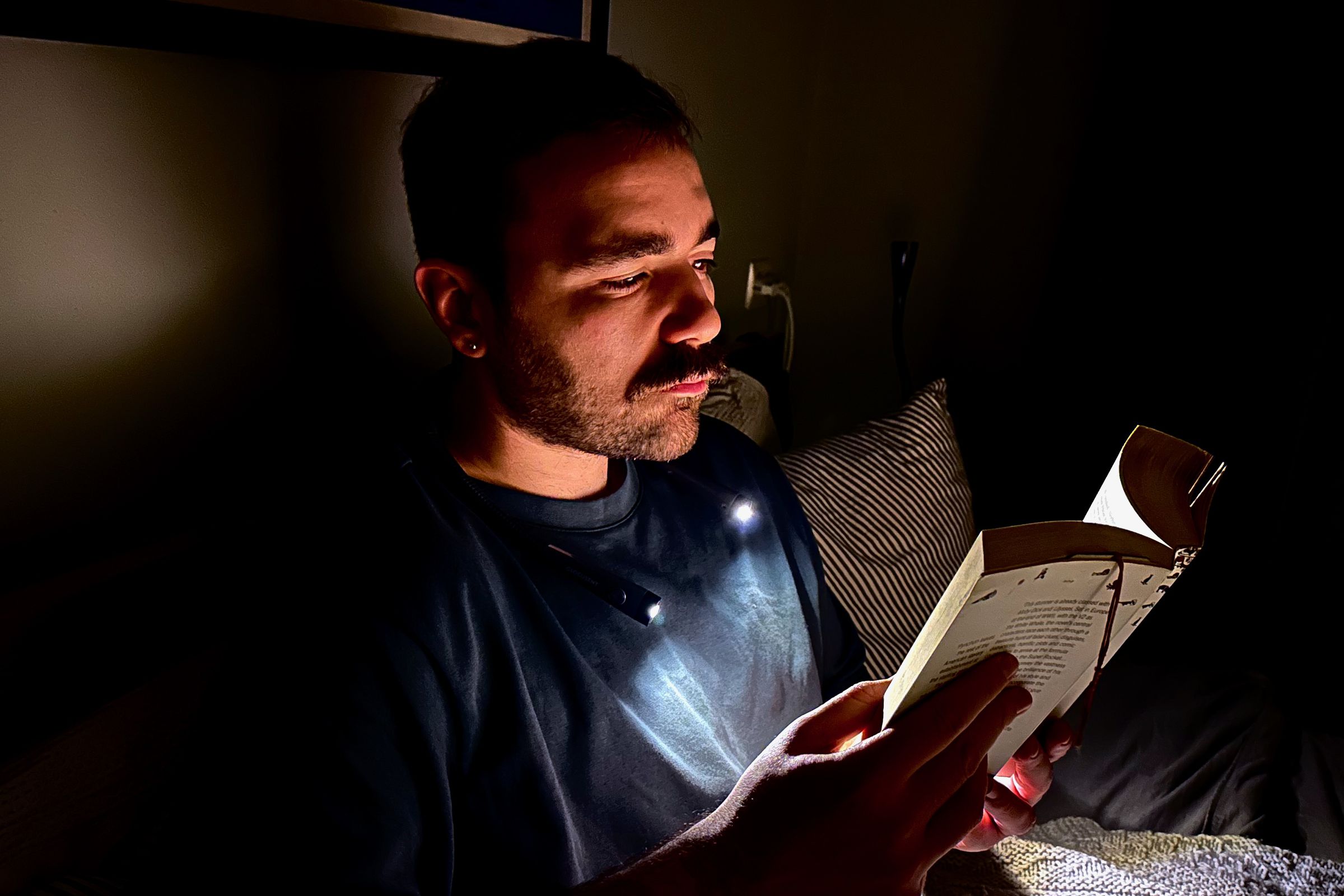 A man in a dark bedroom wears a neck lamp that illuminates just his face and the book he’s holding while sitting upright in bed.