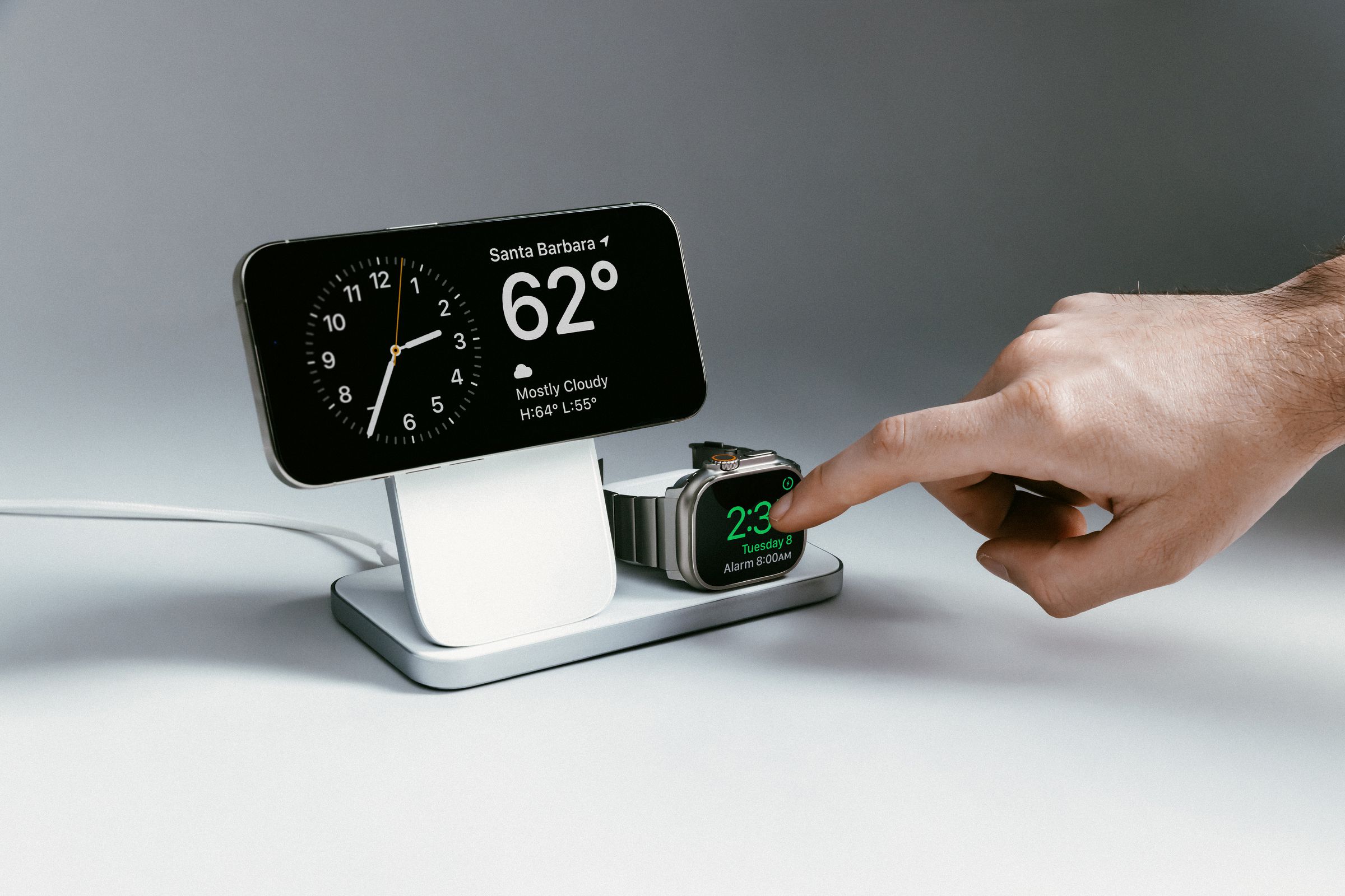 An iPhone and Apple Watch wirelessly charging on a Nomad Stand One Max three-in-one charger. The iPhone is horizontal in StandBy mode and a person is tapping on the Apple Watch screen.