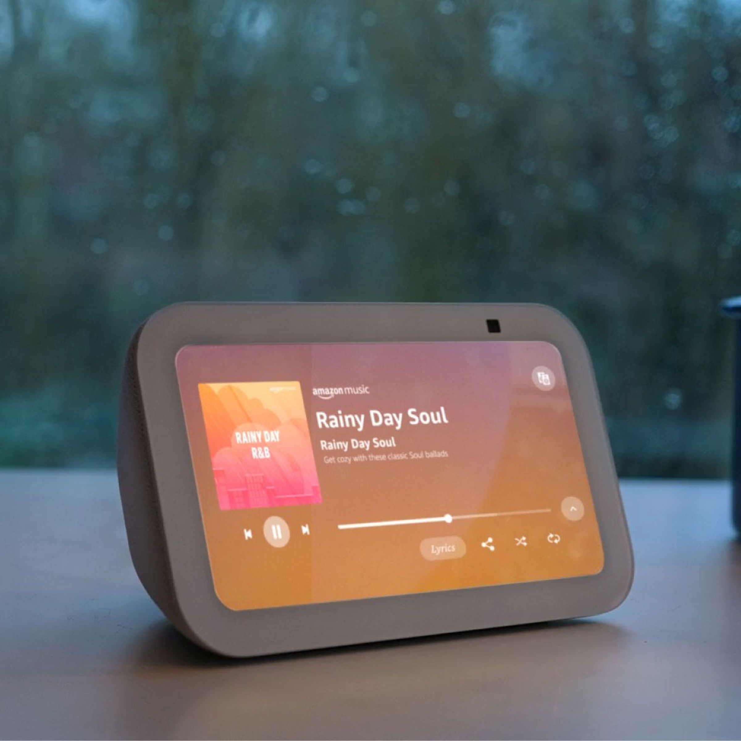 The third-gen Echo Show 5 turned on while on a desk in front of the window during a rainy day.