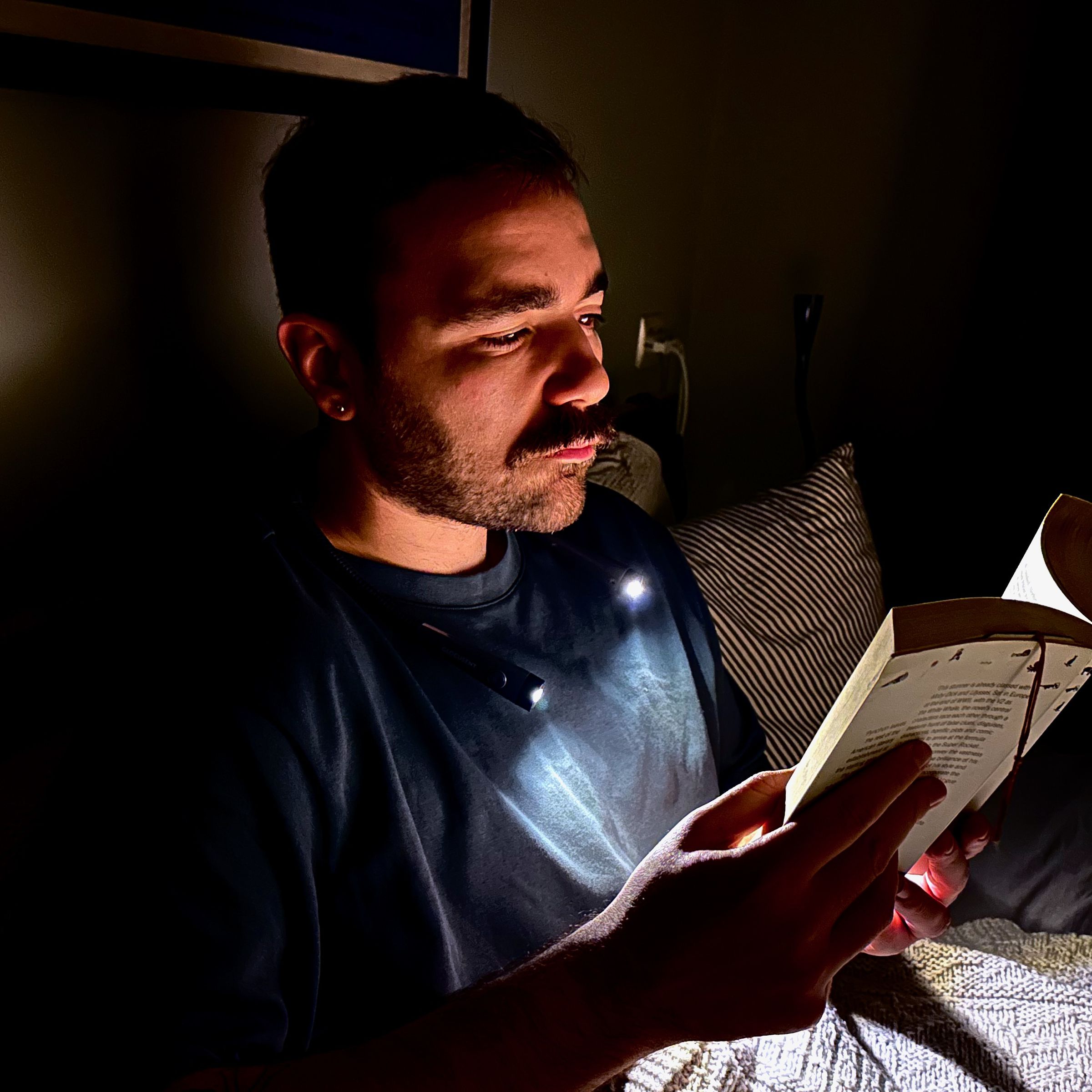 A man in a dark bedroom wears a neck lamp that illuminates just his face and the book he’s holding while sitting upright in bed.