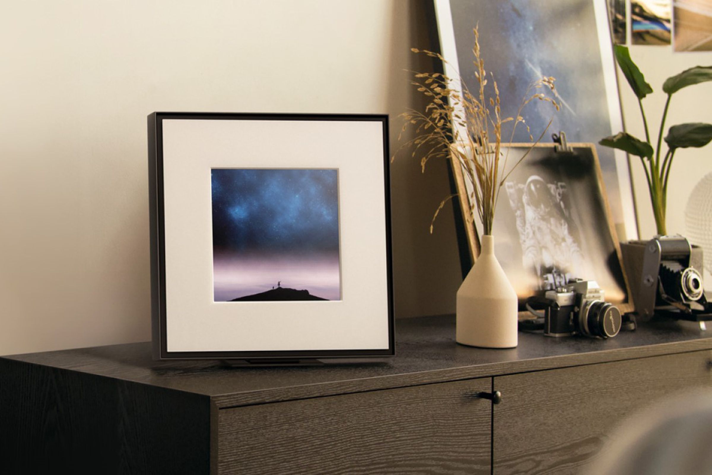 Samsung’s Music Frame speaker sitting on a brown cabinet beside a pair of cameras and some plants.