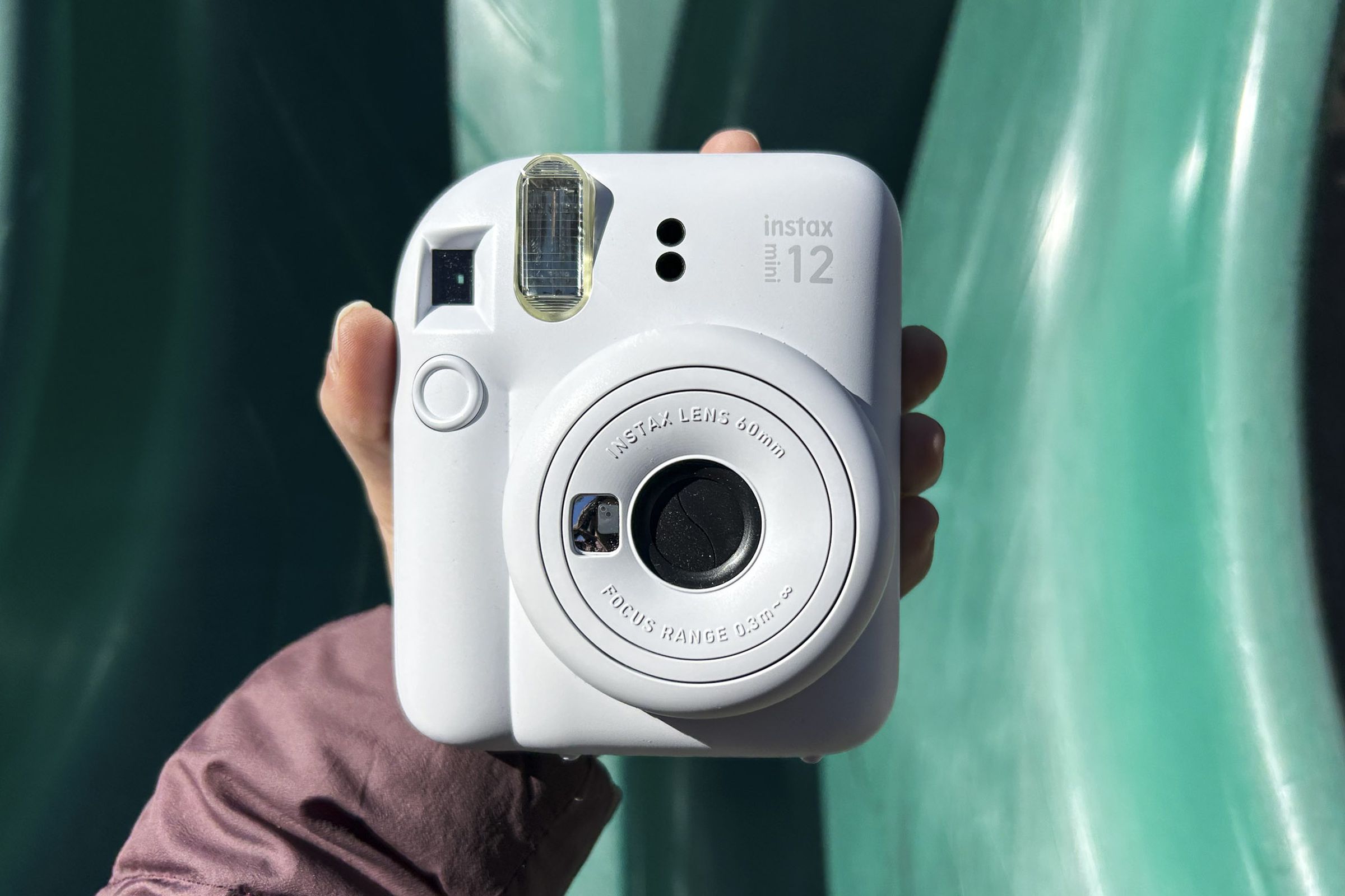 A hand holding up the white Fujifilm Instax Mini 12 against a green slide.