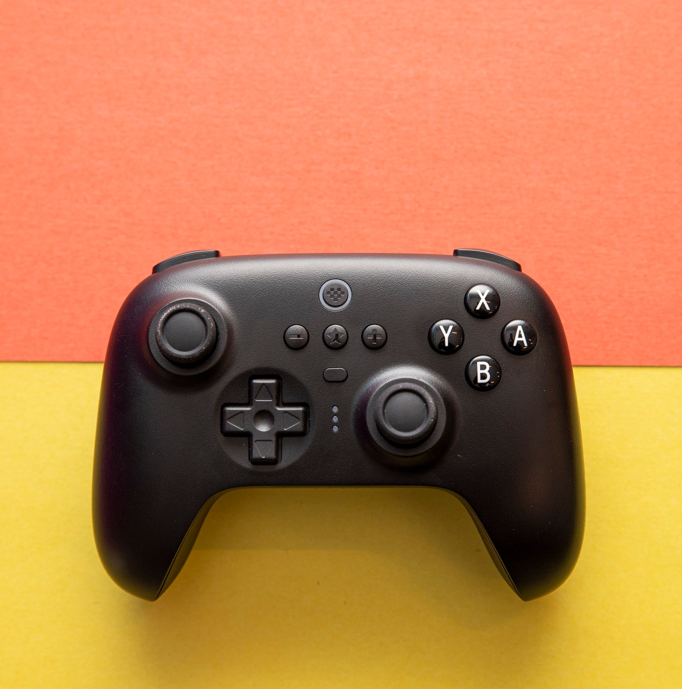 Top-down shot of 8BitDo Ultimate Controller on an orange and yellow background. The controller has an asymmetric joystick design, like an Xbox or Switch Pro controller, and it’s black, with white legends on the A B X Y buttons.