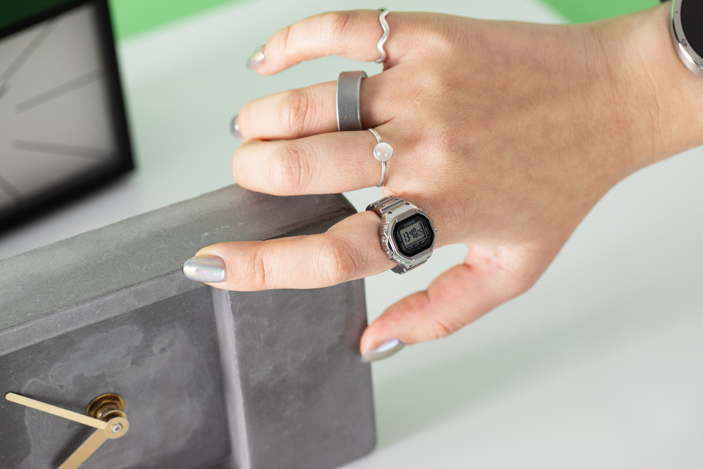 Close-up shot of senior reviewer Victoria Song’s hand with the Casio Ring Watch on her index finger as she reaches for a dark gray stone clock. She has multiple silver rings on and silver holographic nails. There’s a white background.