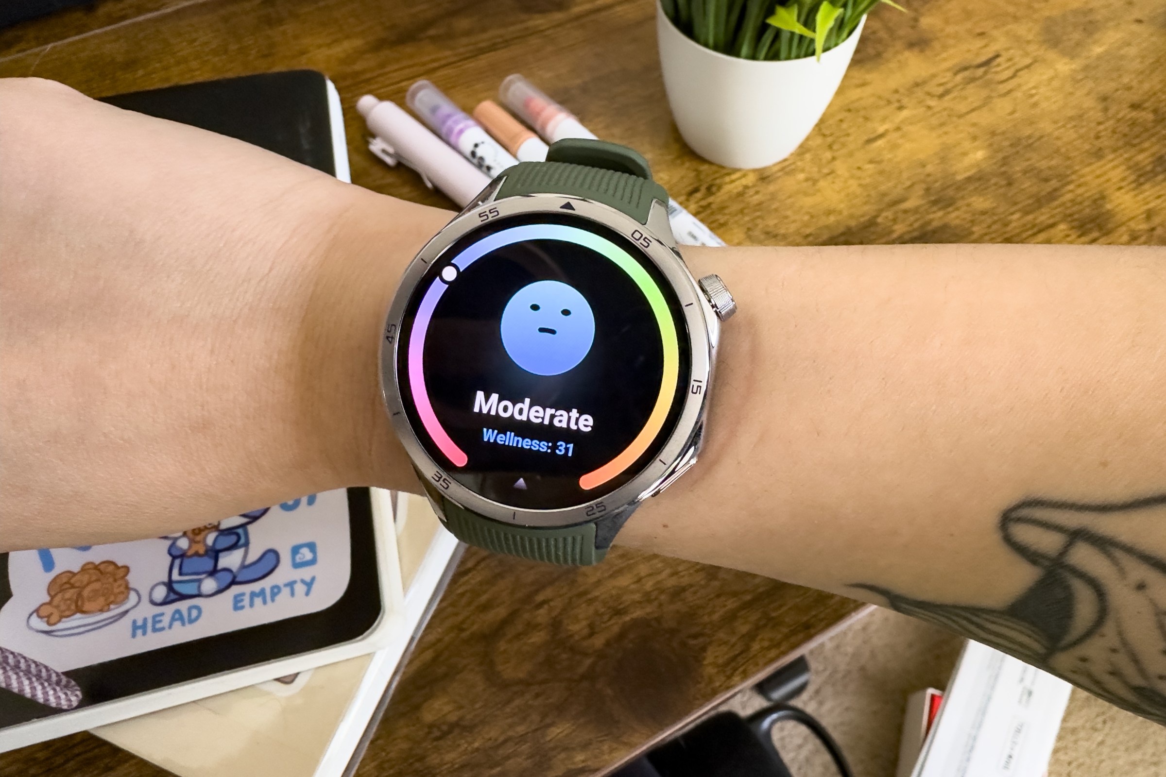 Close up of the OnePlus Watch 3 on senior reviewer Victoria Song’s wrist. It looks slightly large on her smaller wrist. The screen depicts a blank faced blue emoji saying her wellness is moderate. In the background you can see notebooks on top of a desk, pens, and a desk plant.