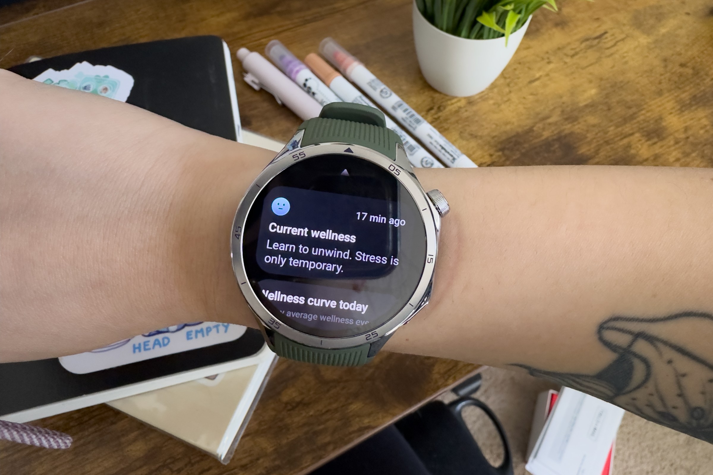 Photo of OnePlus Watch 3 on senior reviewer Victoria Song’s wrist. It shows the 360 Mind and Body Tile depicting Victoria’s current wellness with a blank faced blue emoji. It reads “Current wellness: Learn to unwind. Stress is only temporary.” In the background there is a plant, notebooks, and pens.