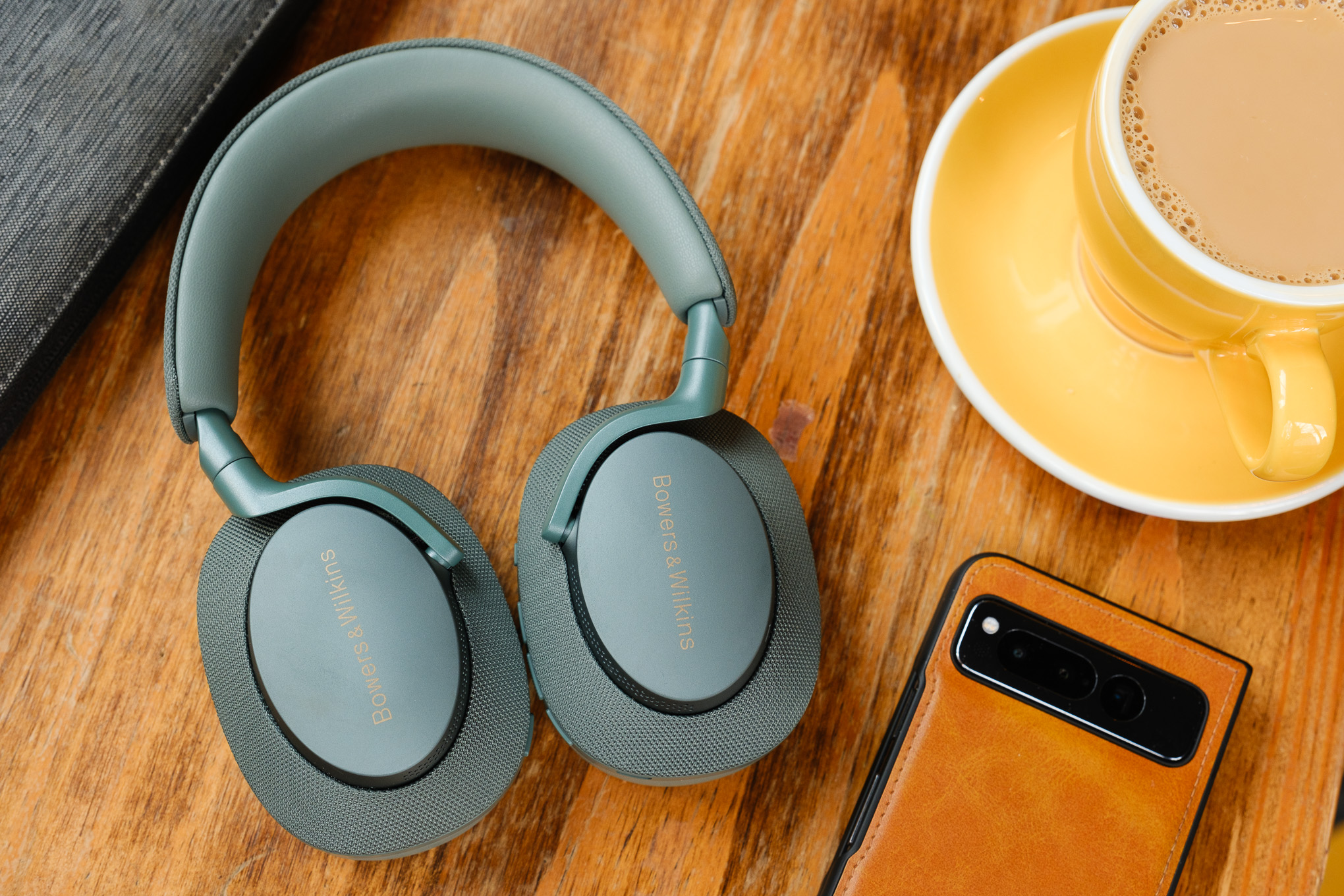 A pair of green Bowers & Wilkins PX7 S2e headphones on a wood table beside a phone and coffee.