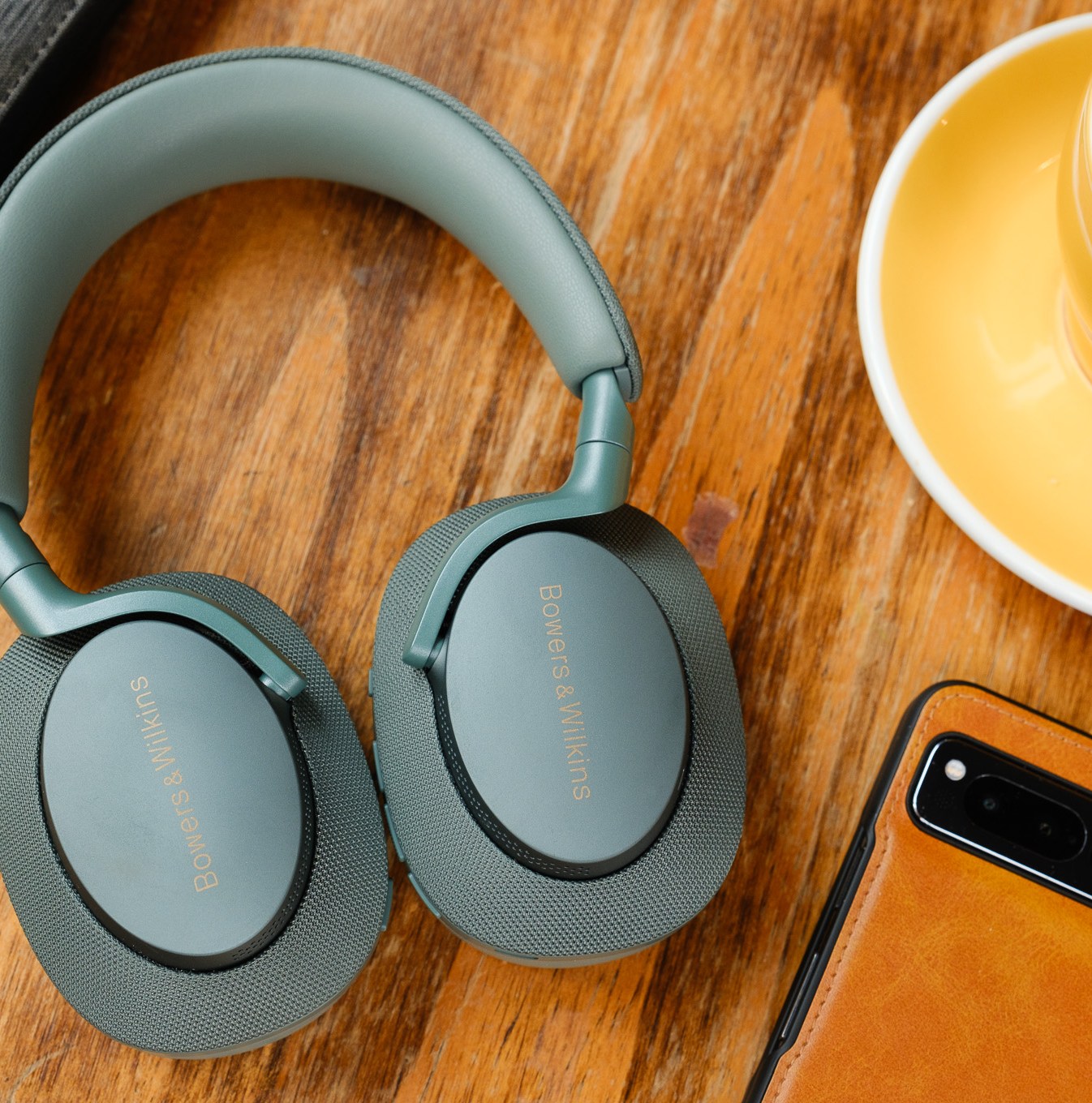 A pair of green Bowers & Wilkins PX7 S2e headphones on a wood table beside a phone and coffee.