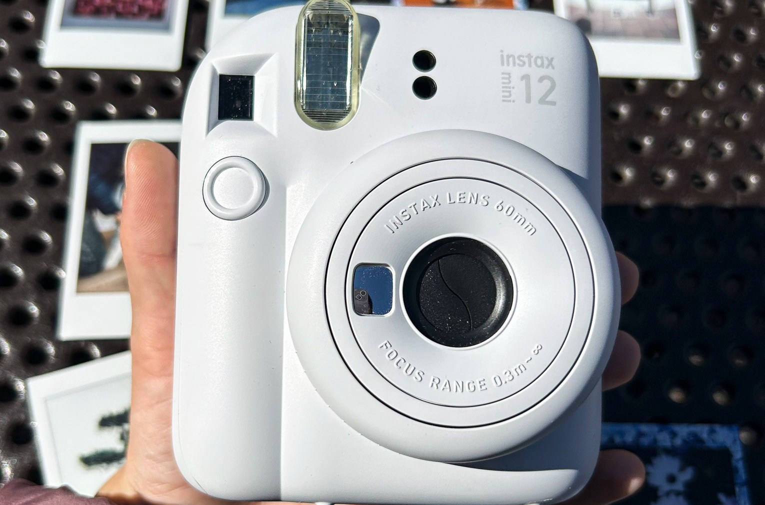 A closeup of Fujifilm’s Instax Mini 12 camera and its lens structure surrounded by photos it printed in the background.