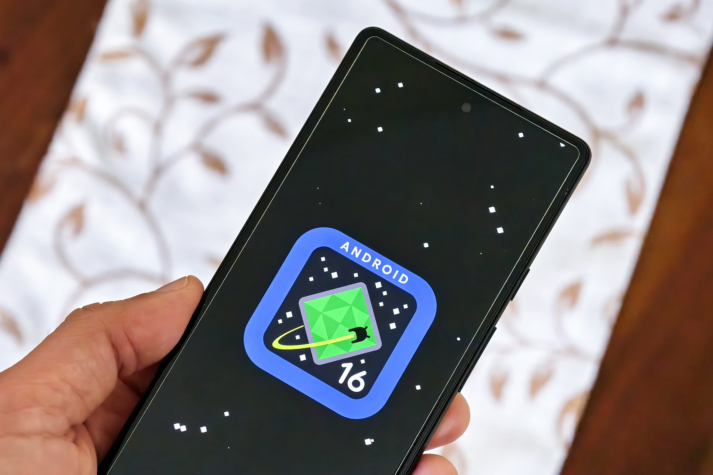 Android 16 logo on Google Pixel 6a held in hand.