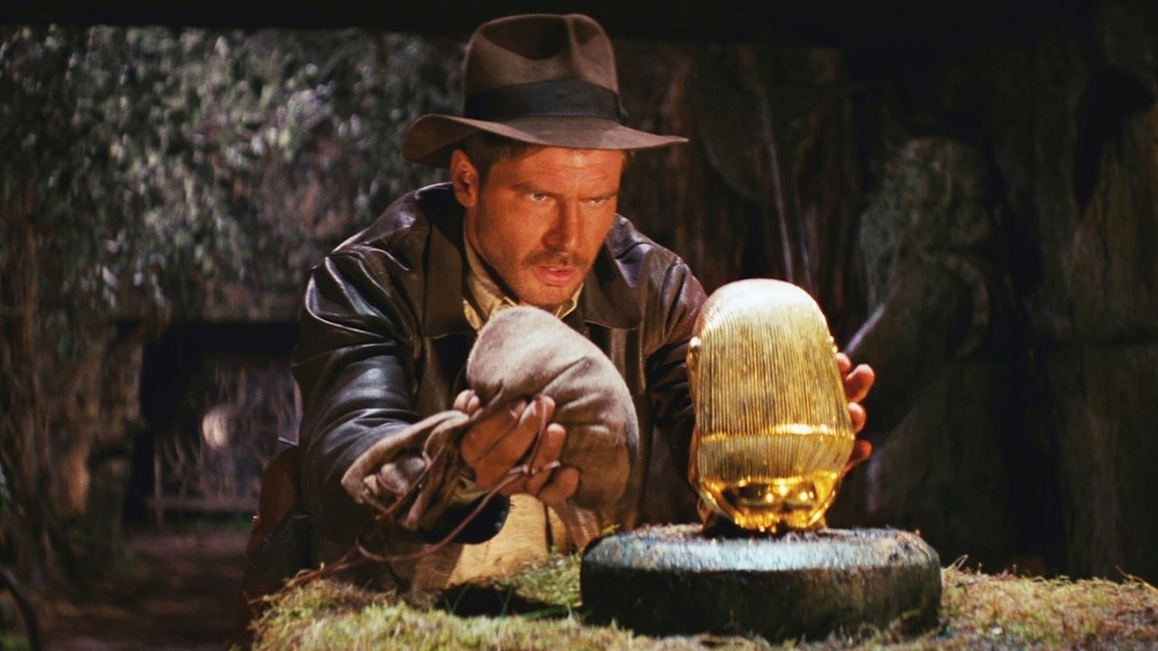Indiana Jones looking at a statue intently while holding a bag in one hand in