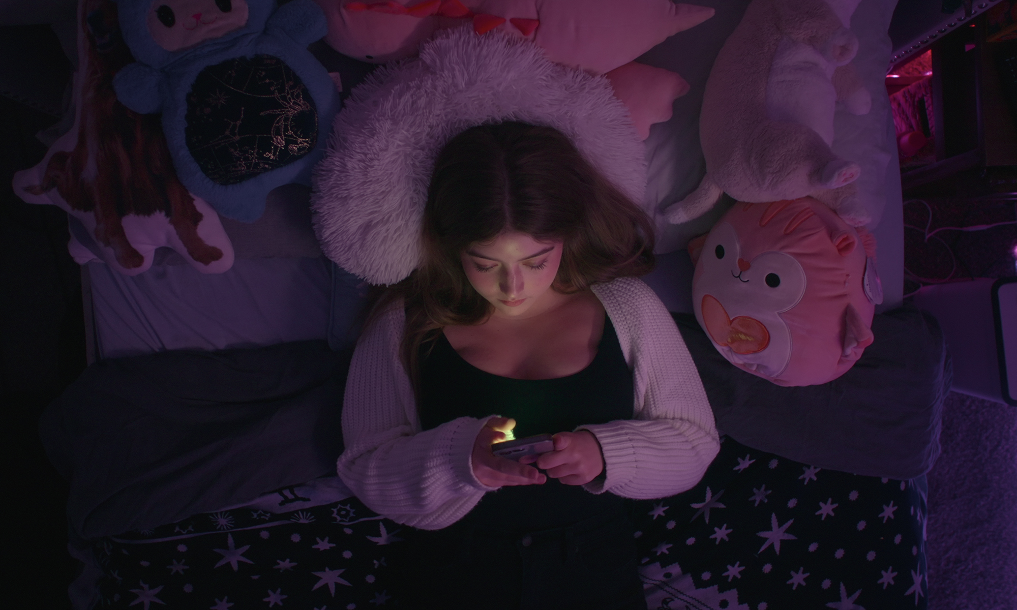 An overhead view of a young teen lying on her bed in the dark looking at her phone from Bad Influence: The Dark Side of Kidfluencing.