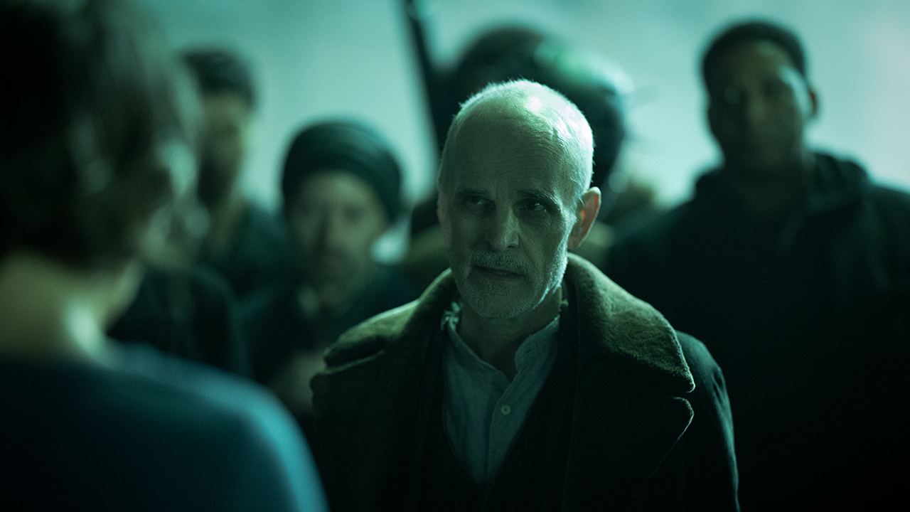 The Croat in a dark setting talking to someone with people behind him in The Walking Dead: Dead City.