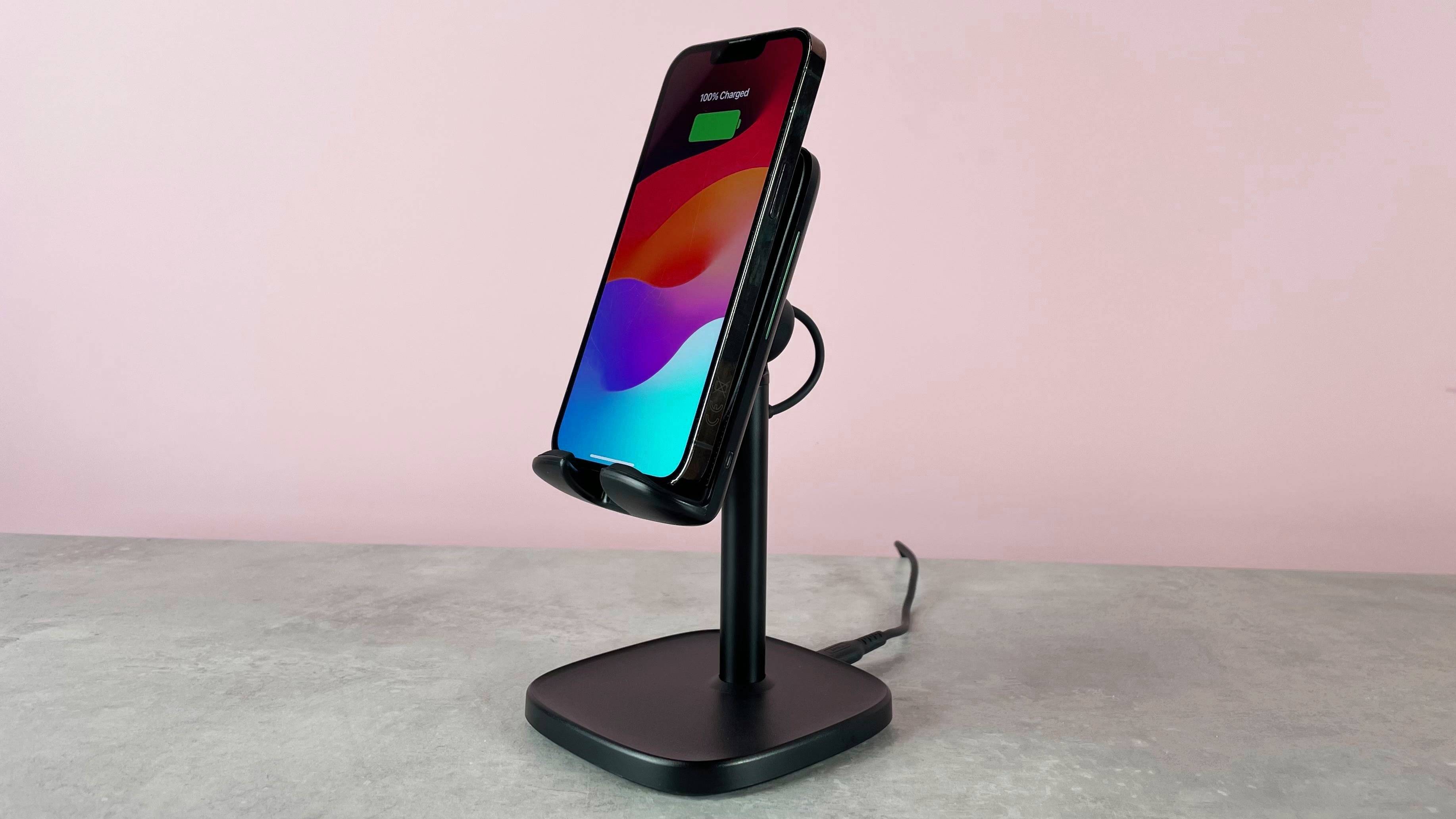 The INIU I212 Adjustable Wireless Charger is pictured sitting on a grey stone surface against a pink background. There is an iPhone on the charger, its screen shows that it is charged to 100%.