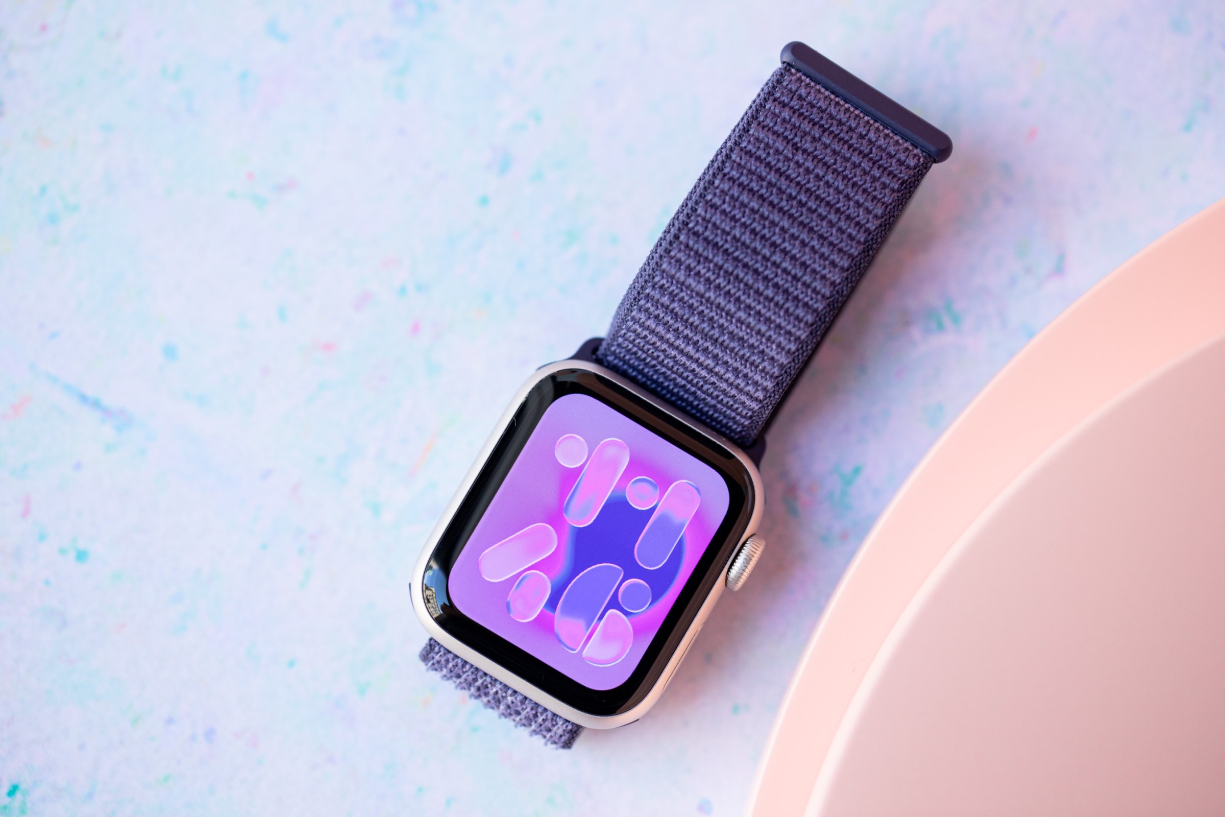 Close up of the Apple Watch SE 3 next to some peach discs on the right and on top of a pastel, blue speckled background.