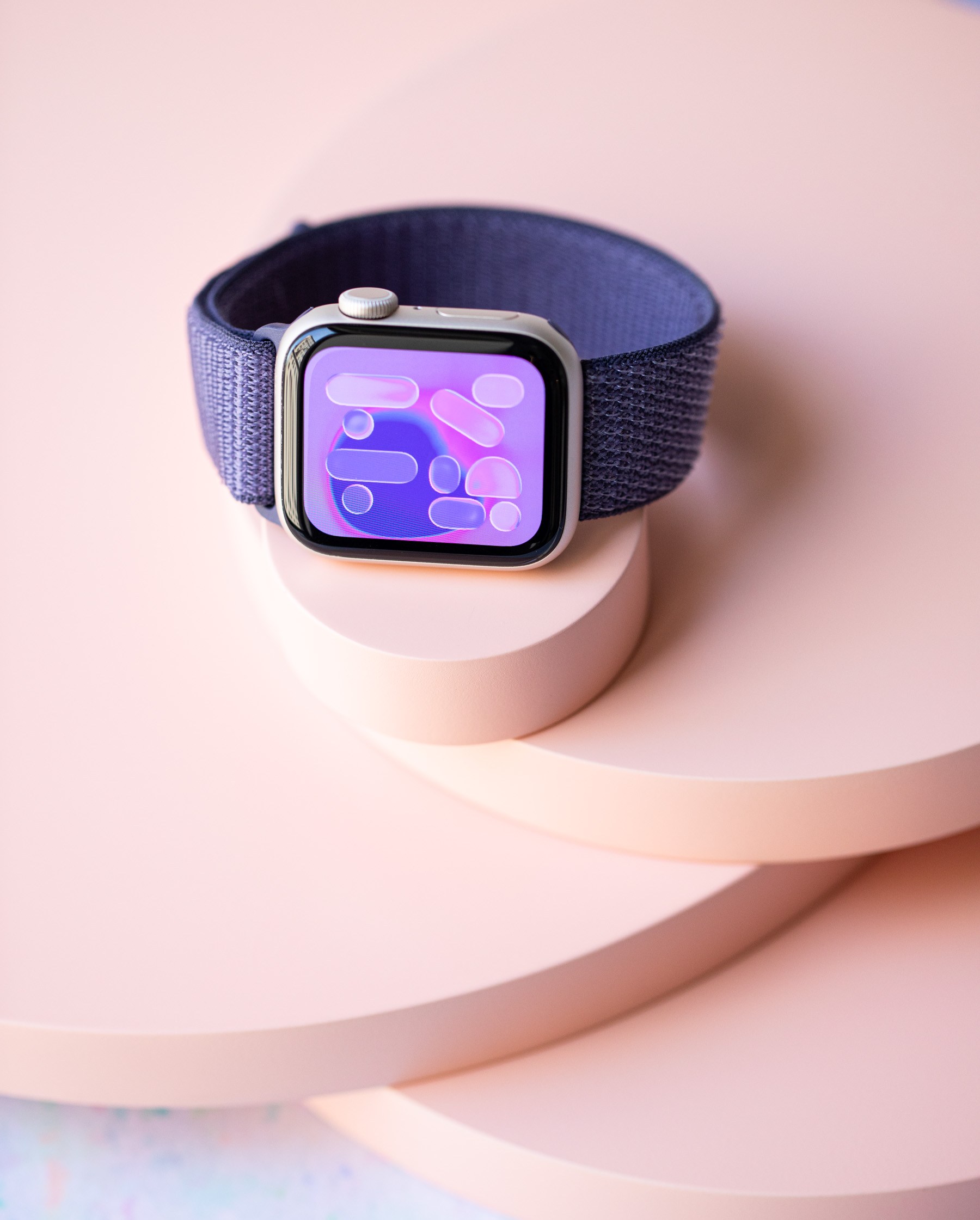 Vertical image of the Apple Watch SE 3 in starlight with a purple nylon band sitting on a series of peach colored discs