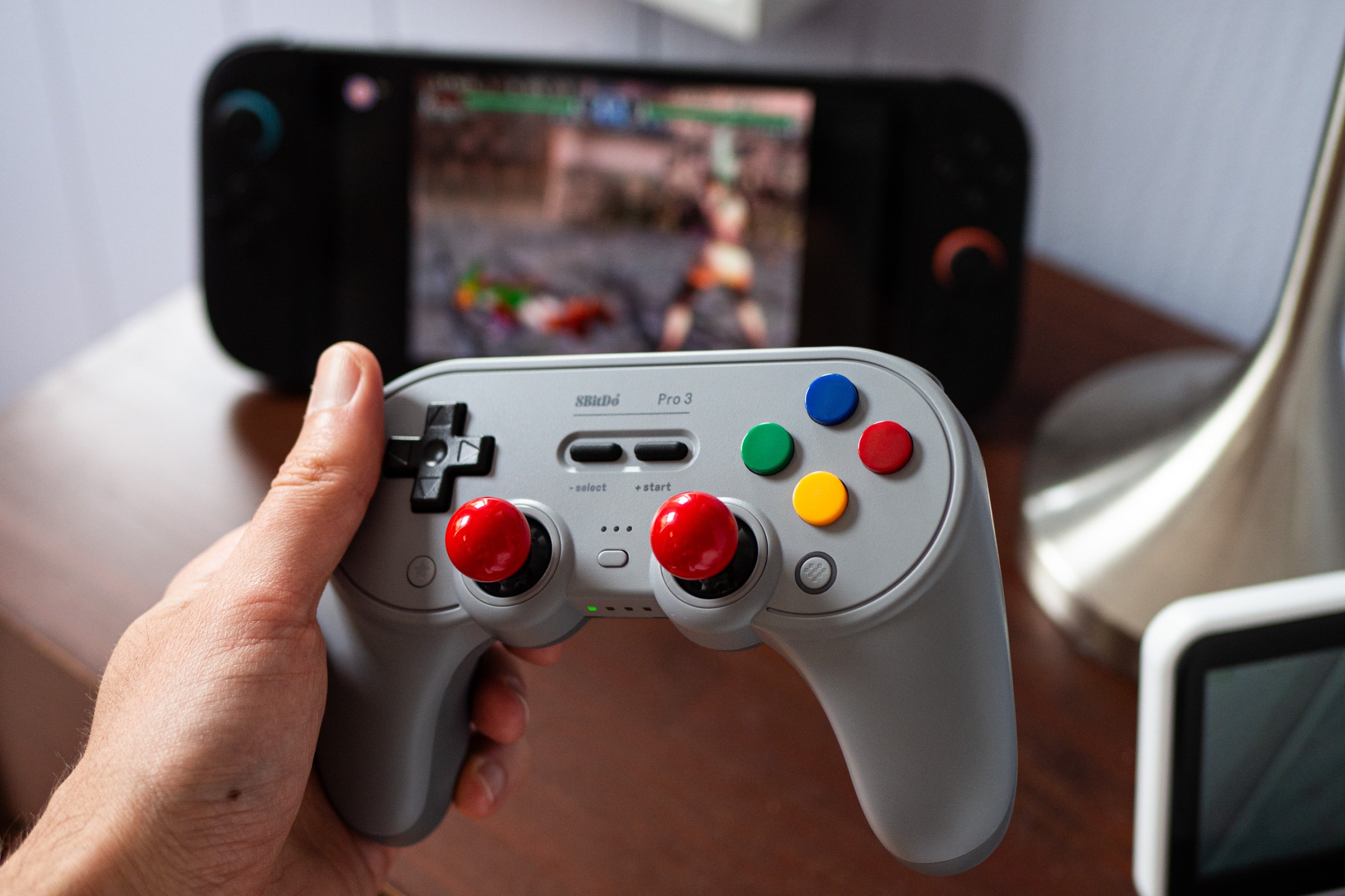 An image showing the author holding the 8BitDo Pro 3 controller with its red arcade stick attachments installed. In the background is a Switch 2 playing Soulcalibur 2.