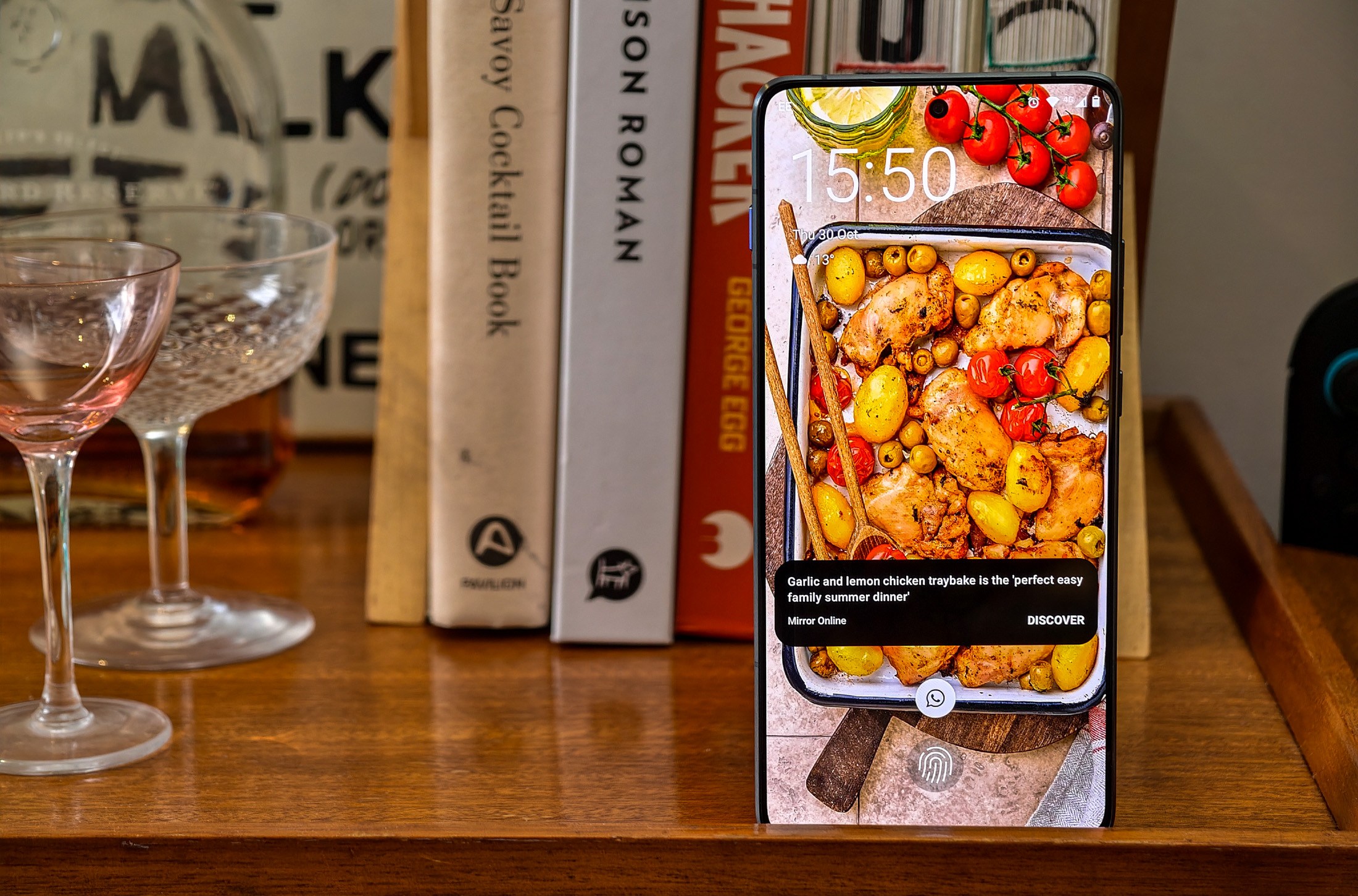 Photo of Motorola Edge 70 standing on a wooden table in front of some cookbooks, showing the live lock screen with an ad for a recipe