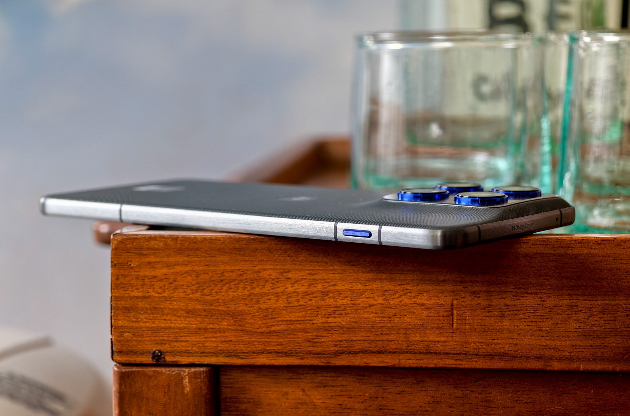 Photo of Motorola Edge 70 lying on a wooden table in front of some glasses, showing how thin the phone is