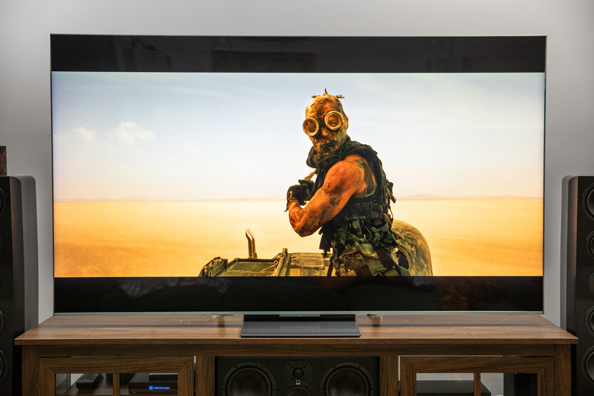 The TCL QM9k on a wooden home theater credenza showing a masked character from Mad Max Fury Road.