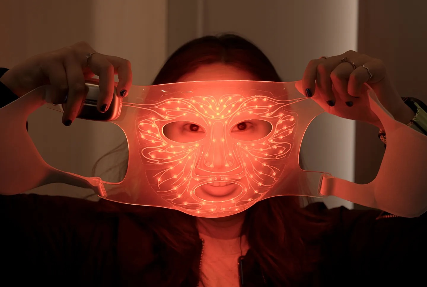 A woman standing behind a sheet mask-style face mask with LED lights across the face