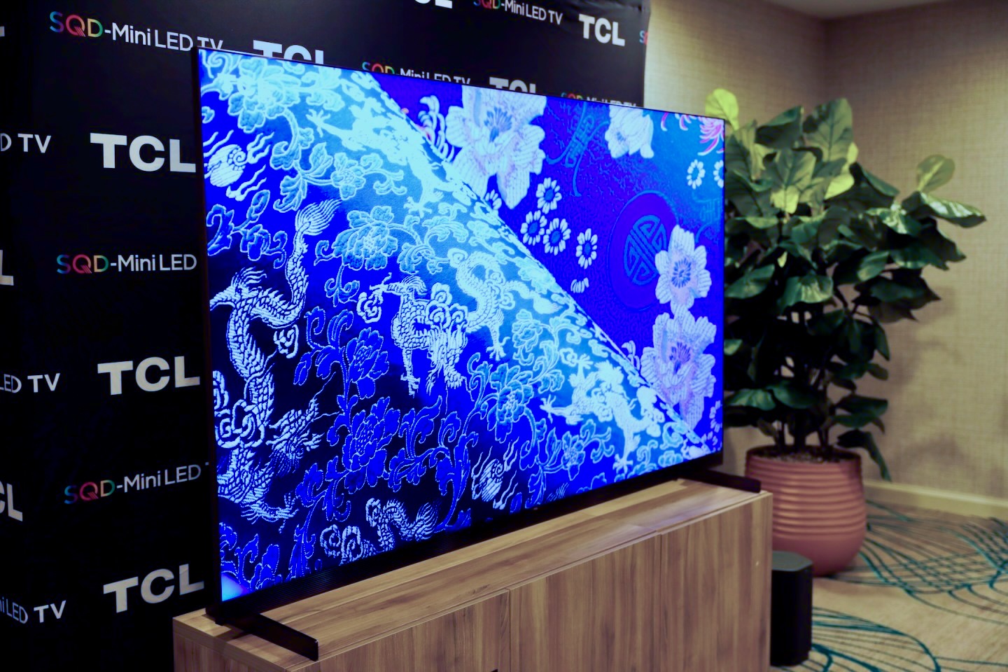 The TCL X11L SQD Mini LED TV on a wooden stand with a TCL-branded backdrop behind it and a potted plant to its side