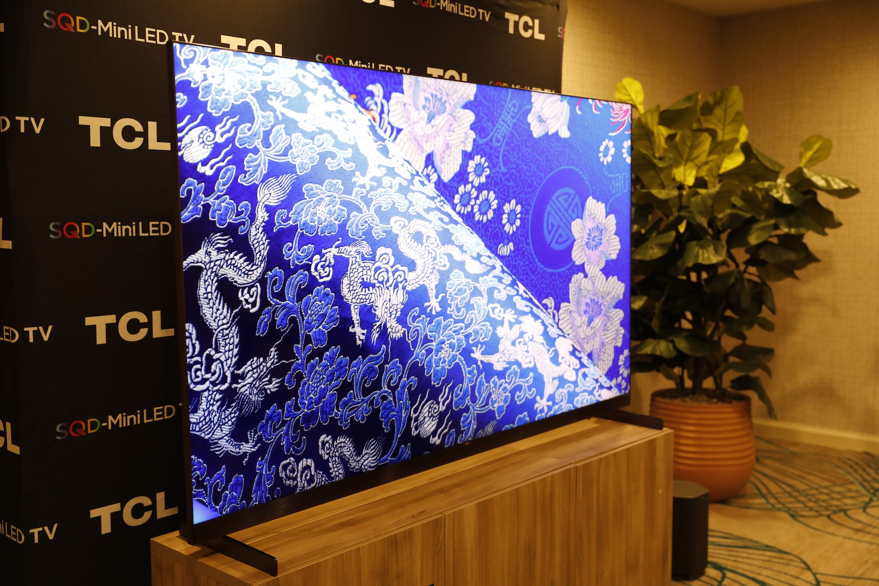 The TCL X11L SQD Mini LED TV on a wooden stand with a TCL-branded backdrop behind it and a potted plant to its side