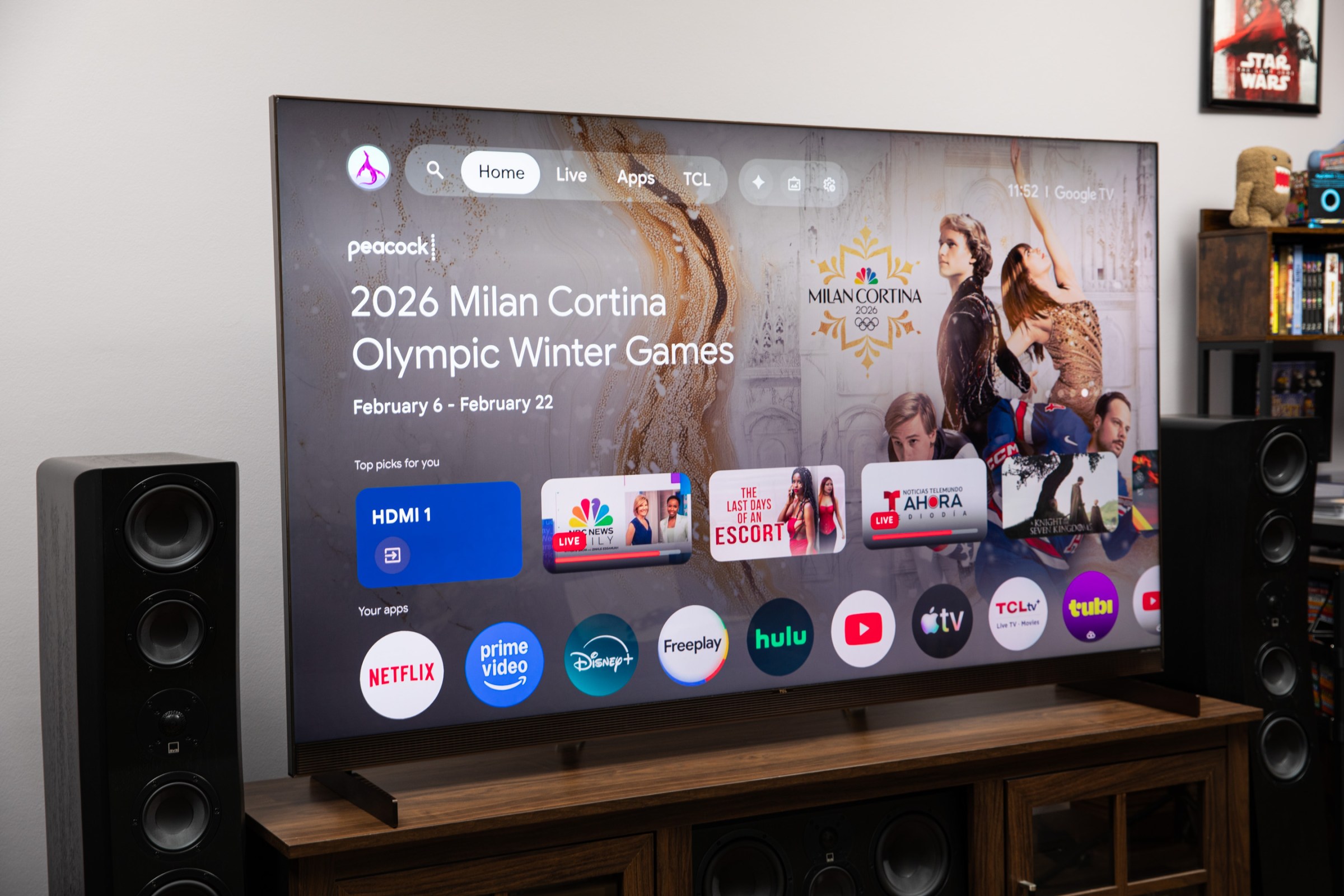 The TCL X11L TV displaying its Google TV OS home page while sitting on a dark wood TV stand.