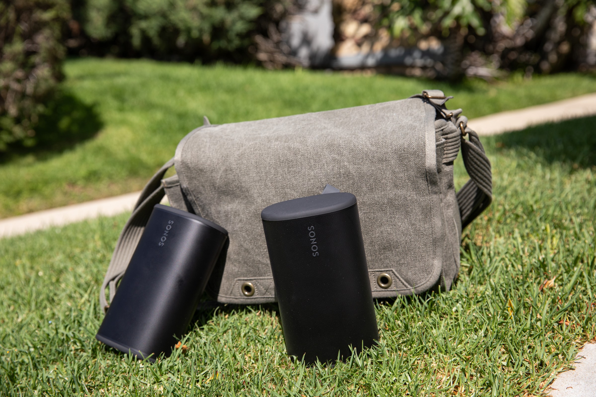 Two Sonos Play speakers in front of a sage colored bag on the grass.