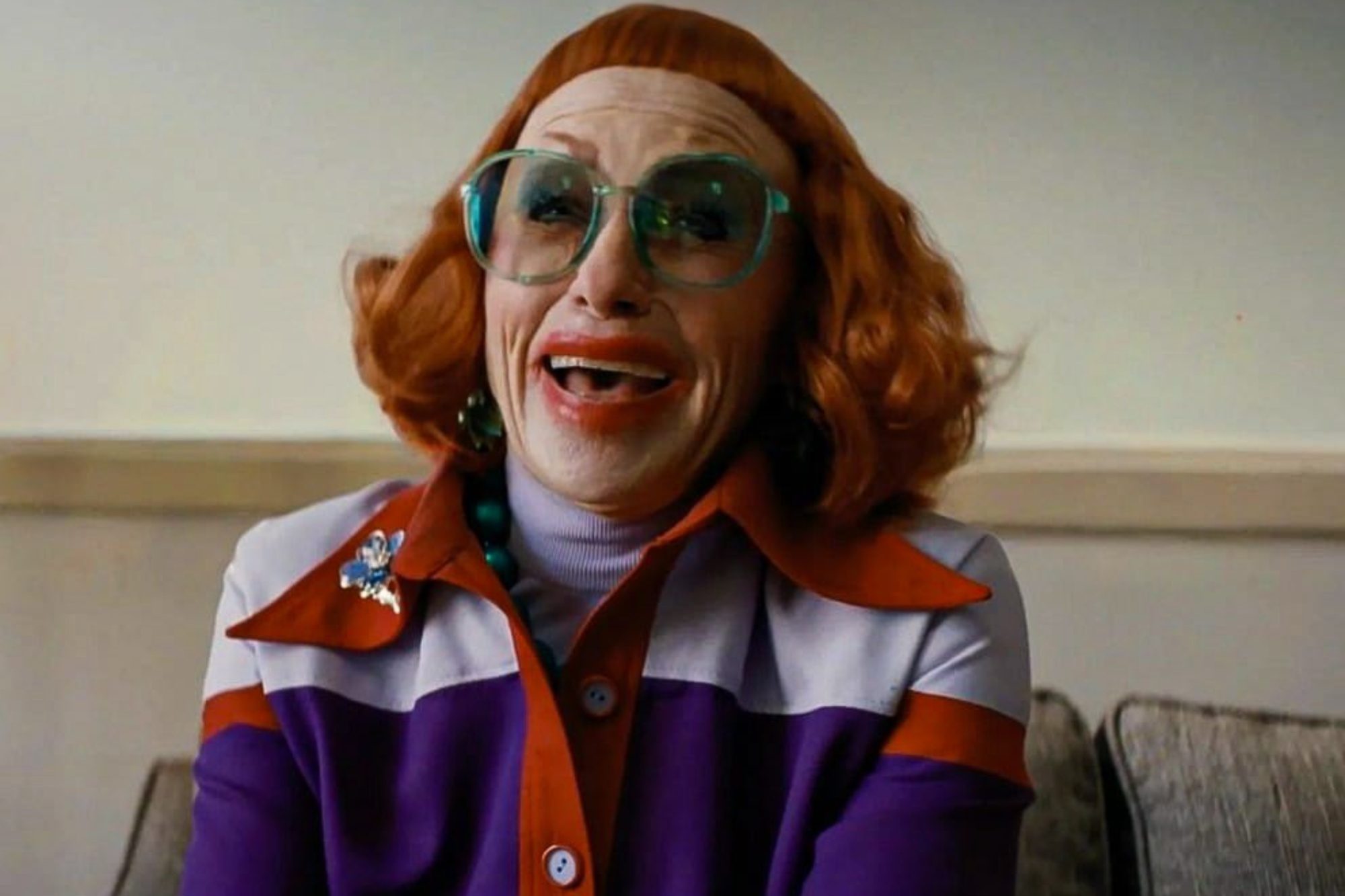 Actor Amy Madigan smiling while wearing big glasses and a red wig as Aunt Gladys in Zach Cregger's horror film, Weapons.