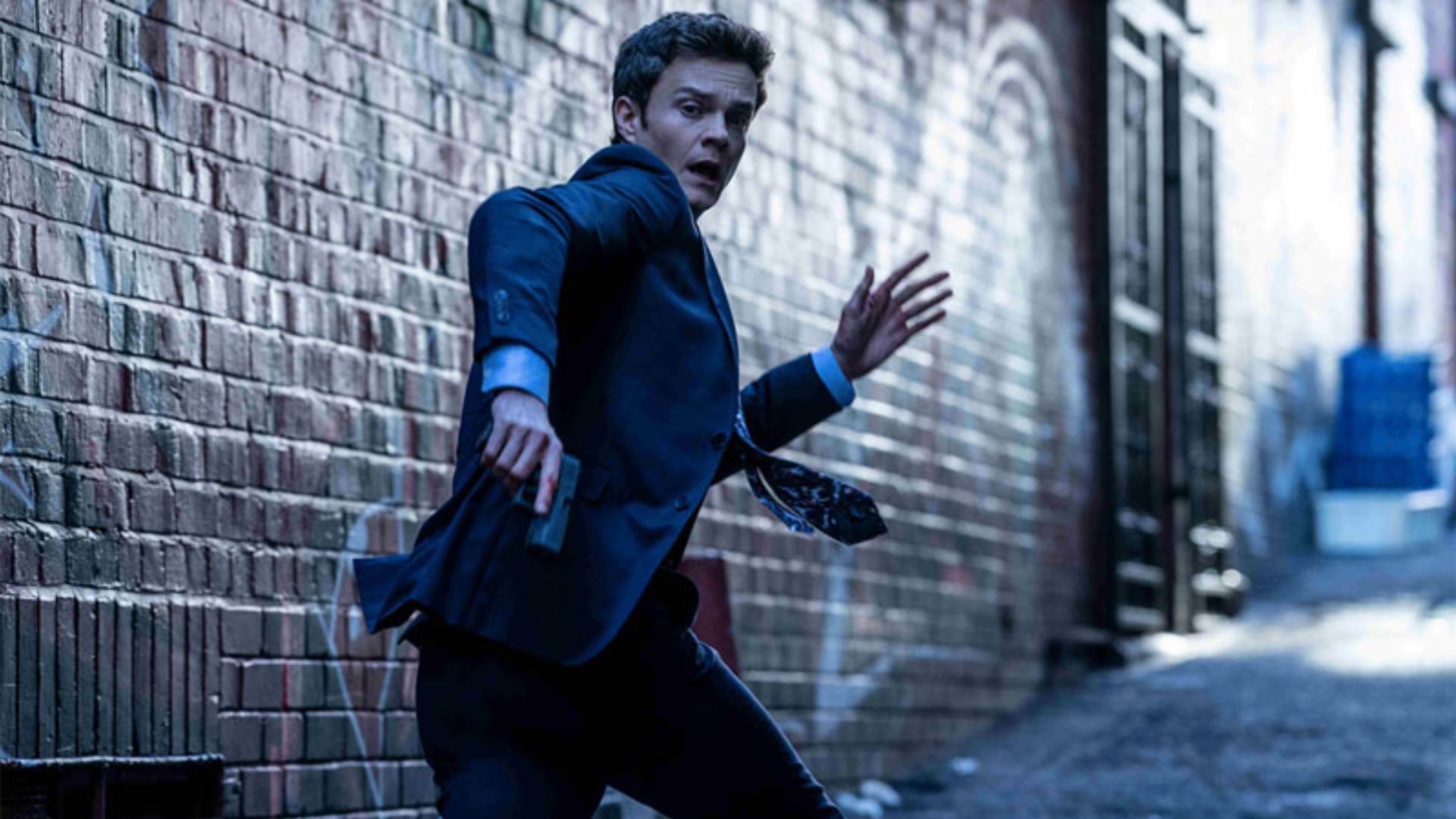 Jack Quaid runs into an alley with a gun in his hand in Novocaine.