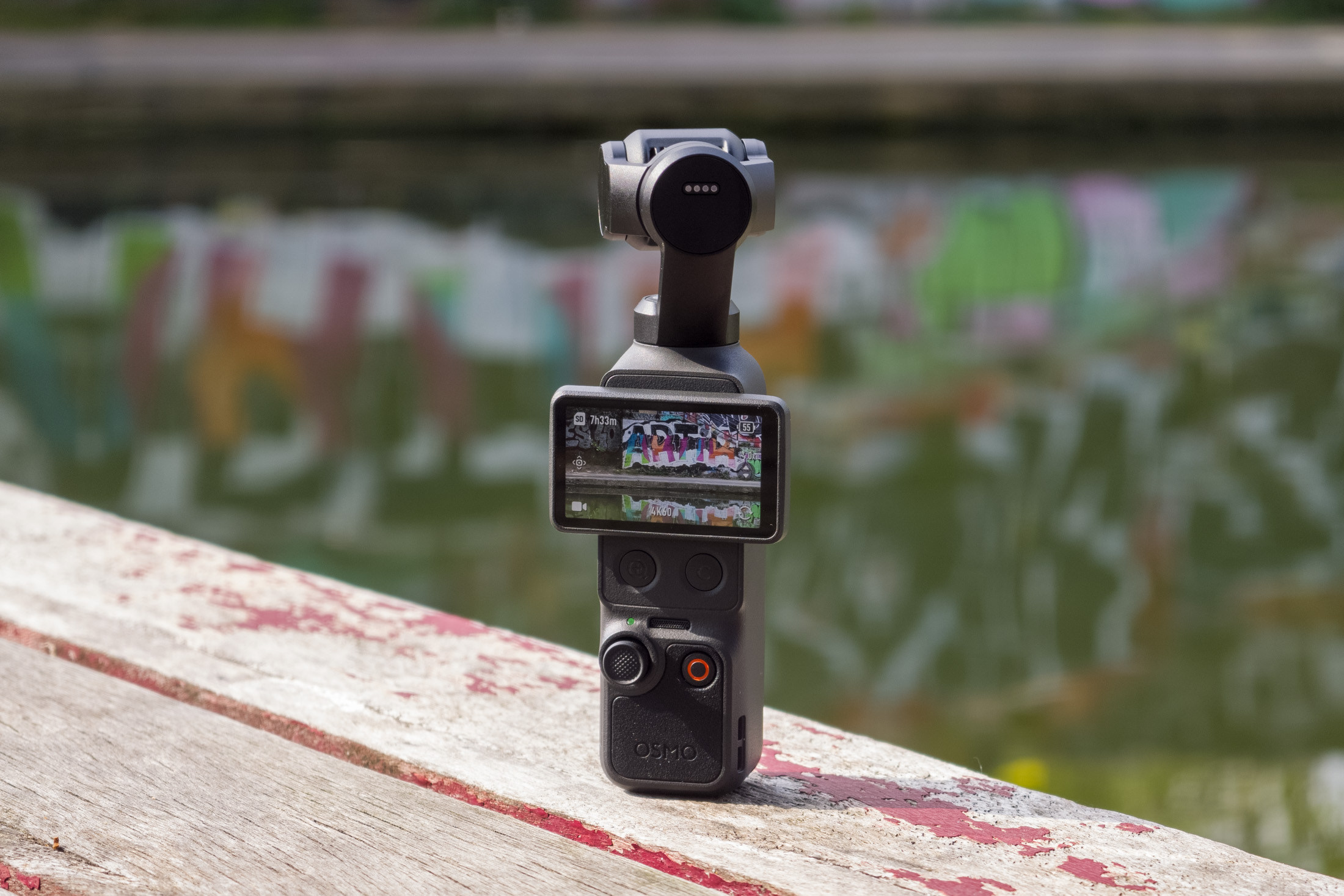 Photo of the DJI Osmo Pocket 4 standing on a wooden table in front of a canal, showing the screen with the camera view