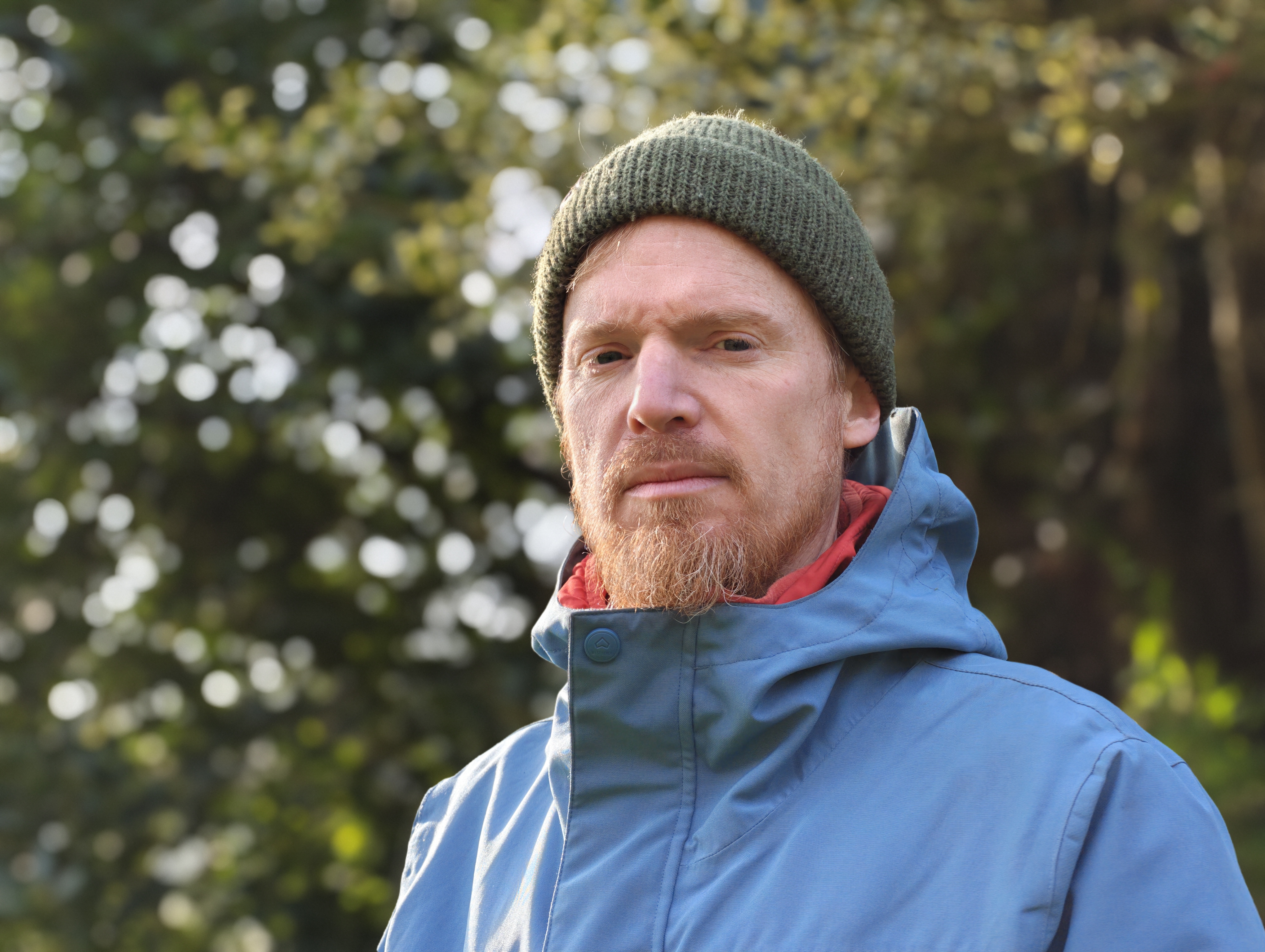 Portrait of a man in a blue coat and green beanie, with dappled light in the background