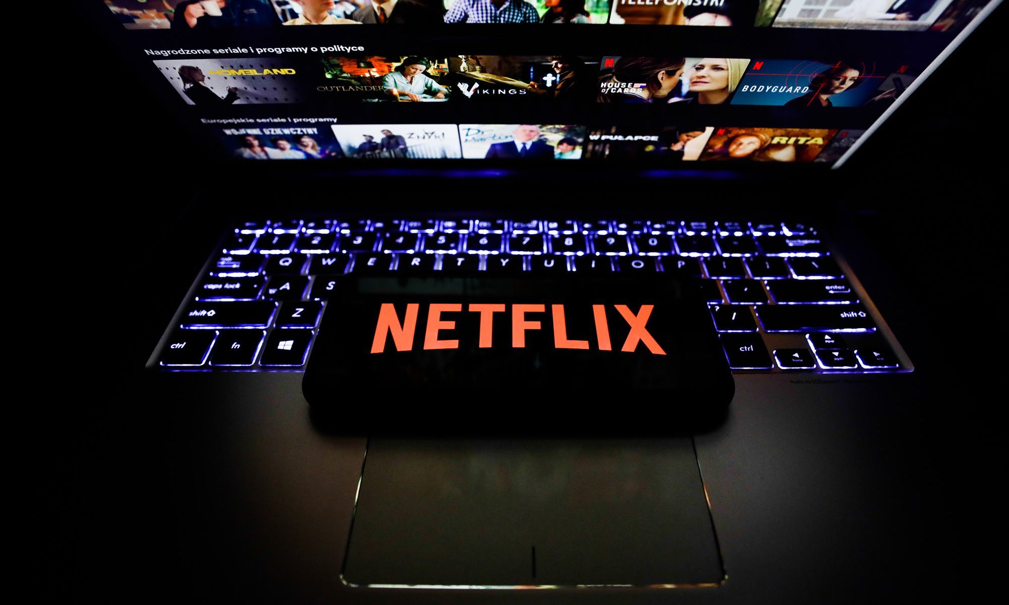Netflix logo is seen displayed on a phone screen while the desktop app is shown on a laptop