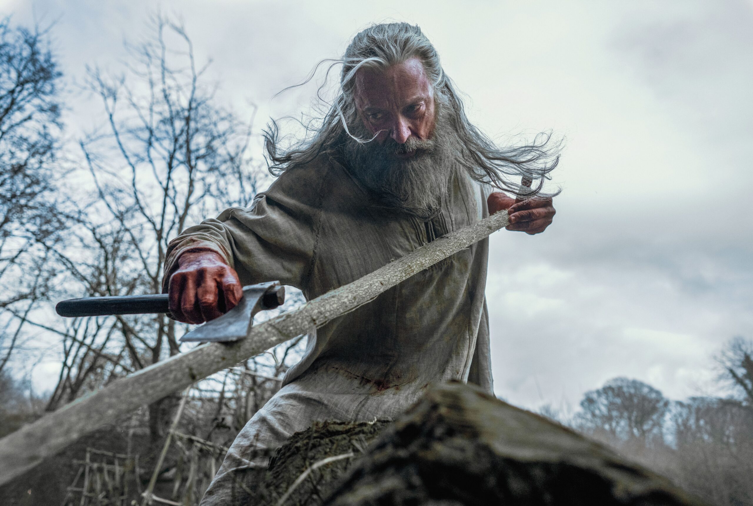 Hugh Jackman sculpting a wooden bow with an ax in the A24 film, The Death of Robin Hood.