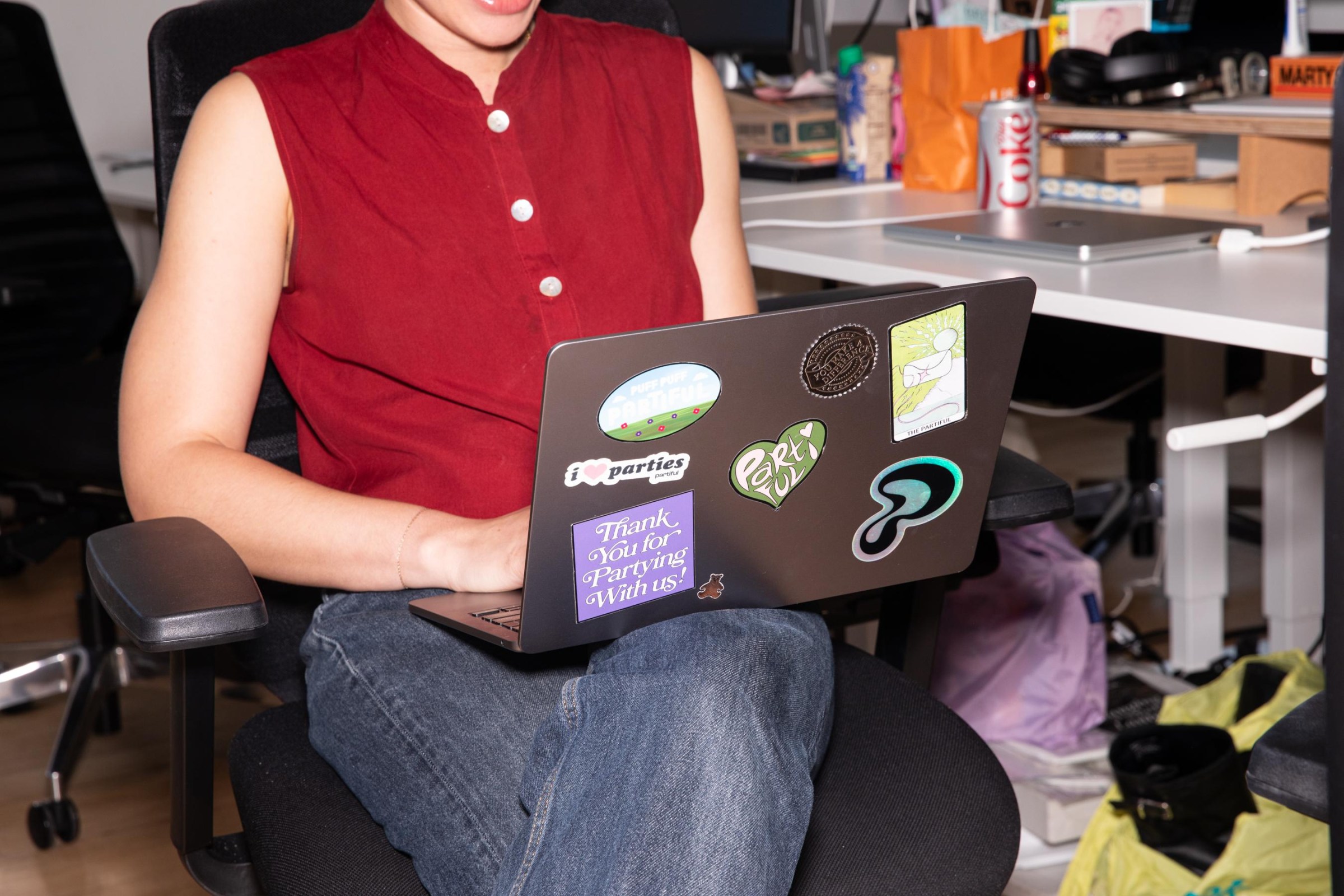 A person in a red sleeveless button down shirt and jeans sits in an office chair with a laptop on their lap which has partiful logo stickers and others that read, “thank you for partying with us” and “I (heart) parties.“