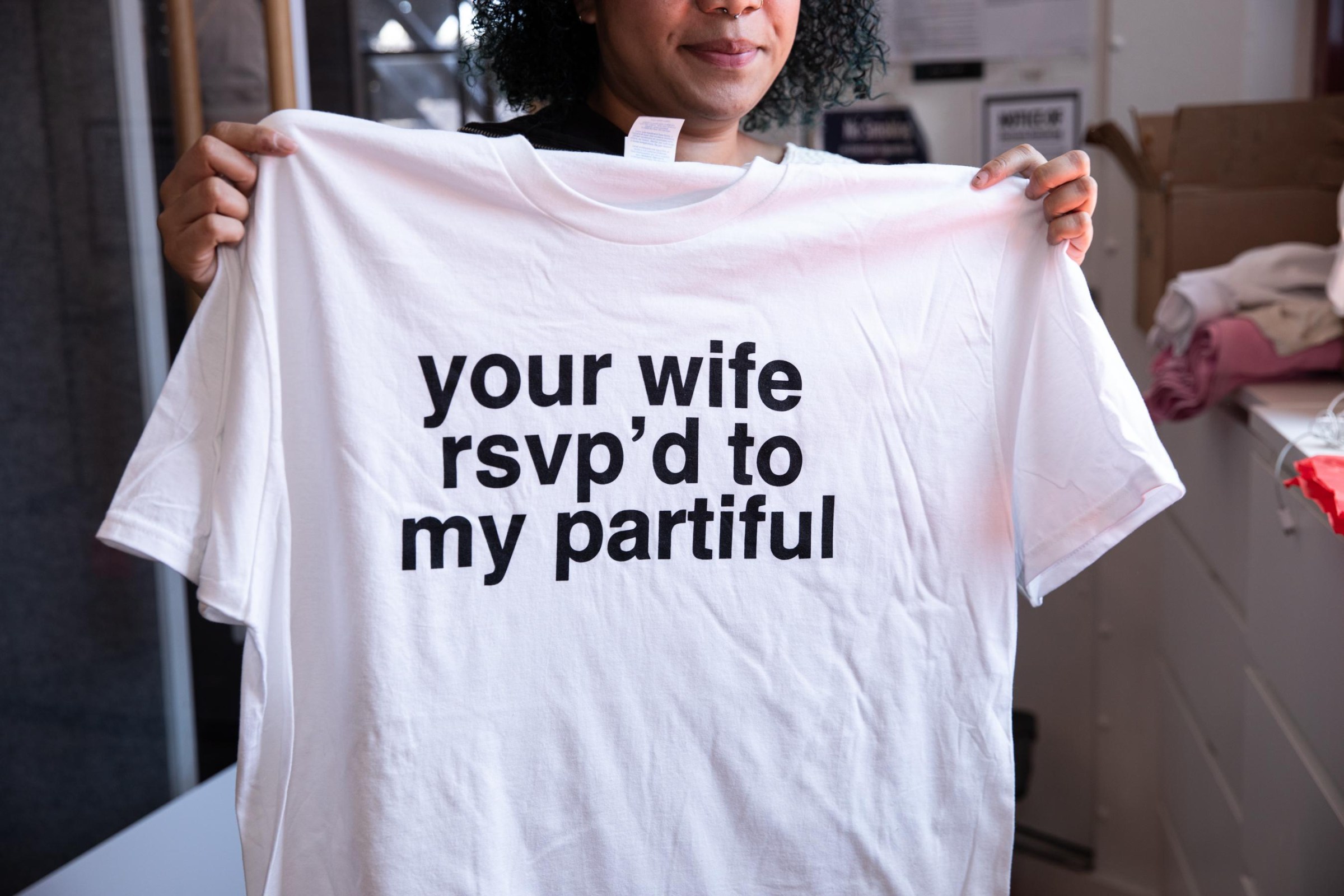 a person holds a white T-shirt that says ‘your wife rsvp’ed to my partiful’ in bold black font.