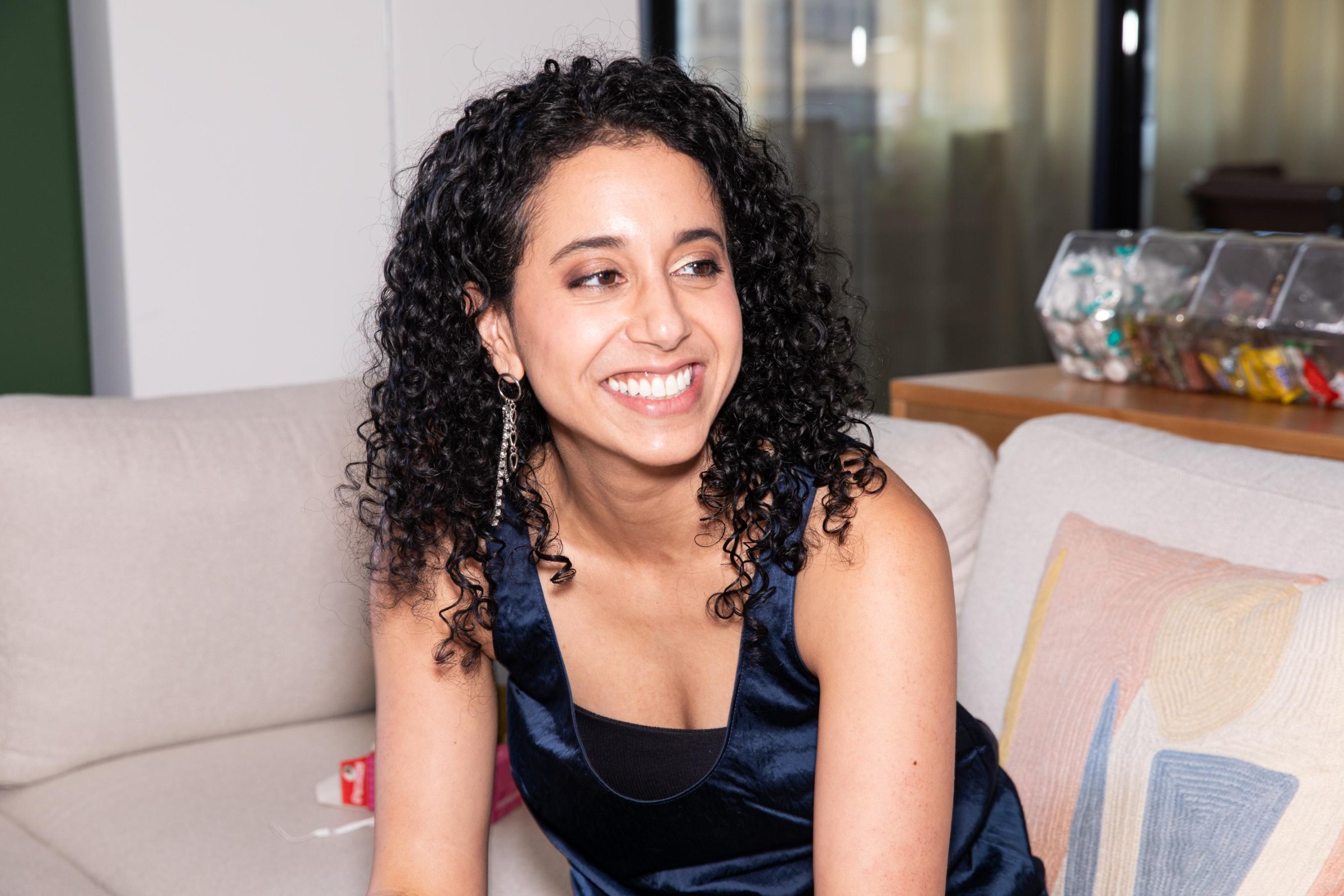 A young woman with curly dark brown hair wearing a sleeveless blue top looks to the side and smiles while sitting on a couch.