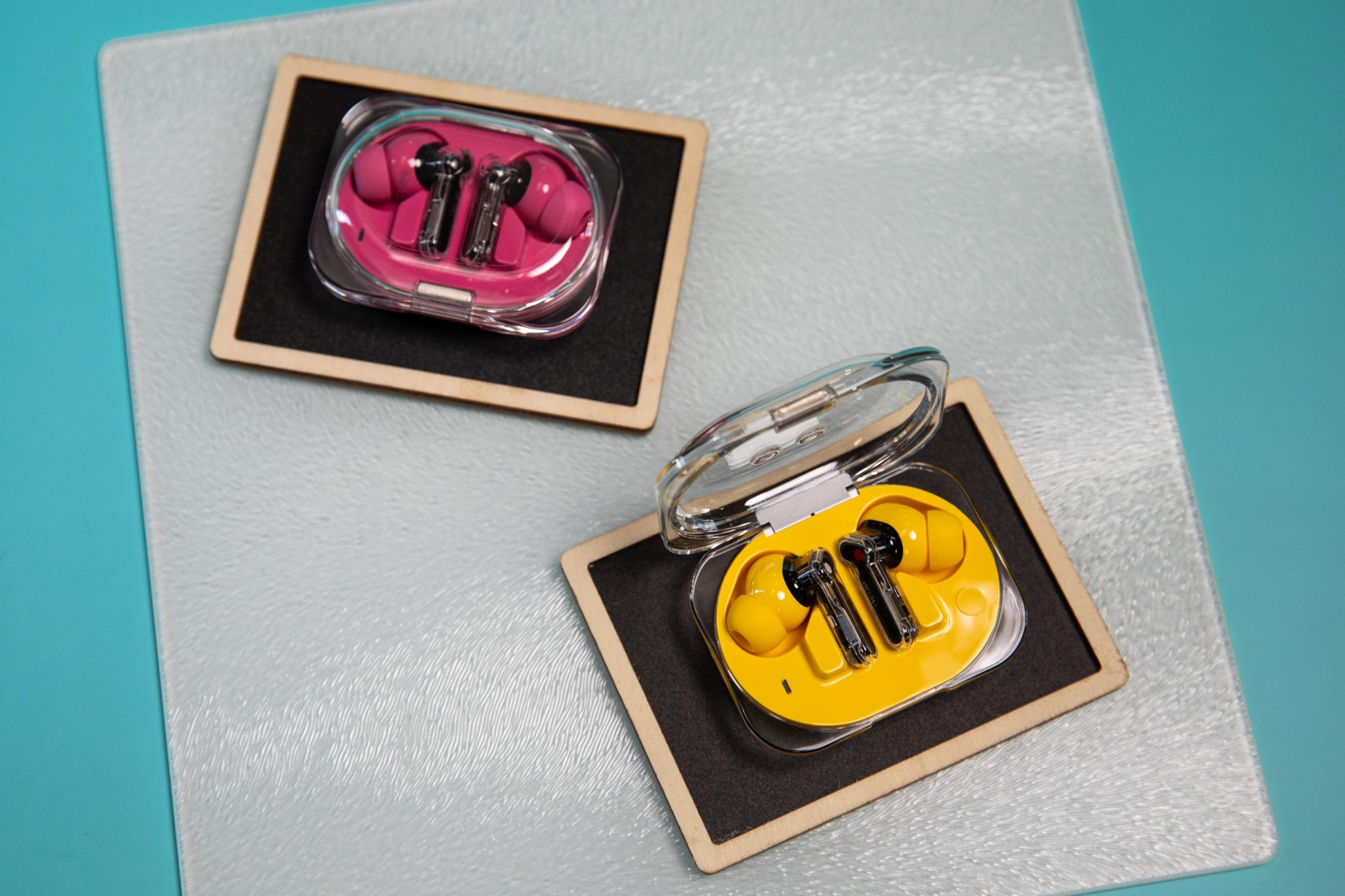 The Nothing Ear 3A earbuds in both yellow and pink on a textured surface and teal background.