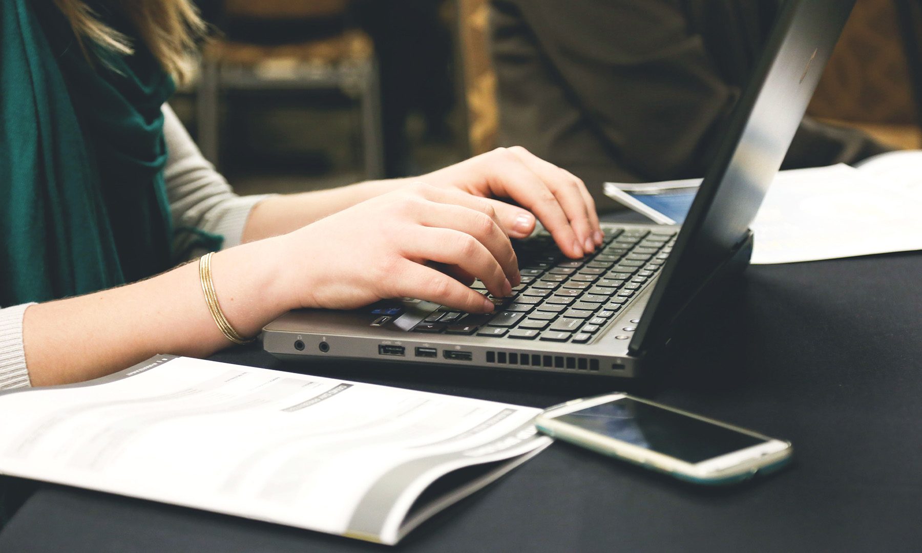Adult woman typing on a laptop at a desk with a smartphone and documents
