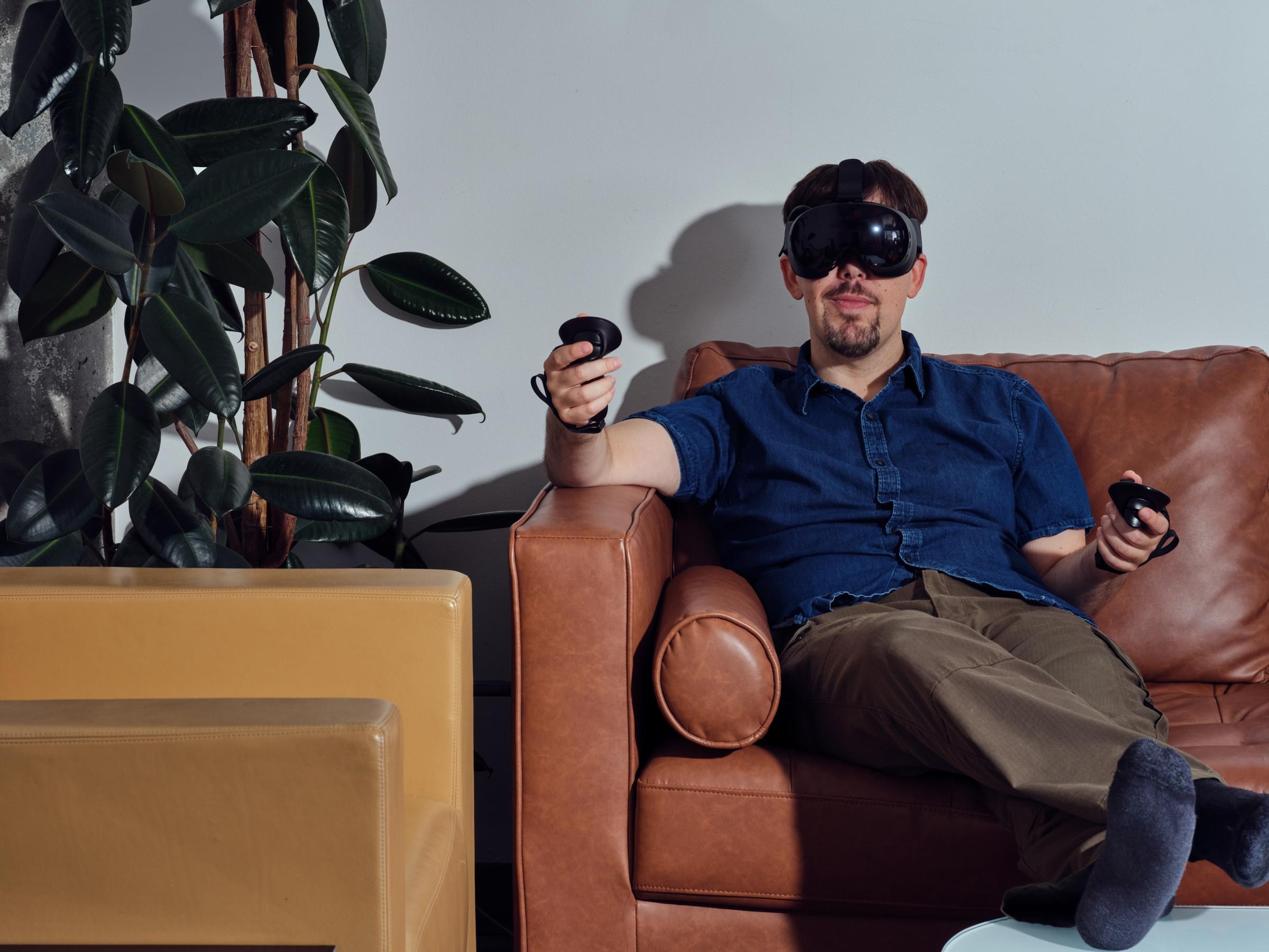 Sean lounges on a couch facing the camera while inside the Steam Frame, with one wand-holding arm propped up on an armrest and the other against the rear cushion. A rubbery leaf plant is visible to his right.